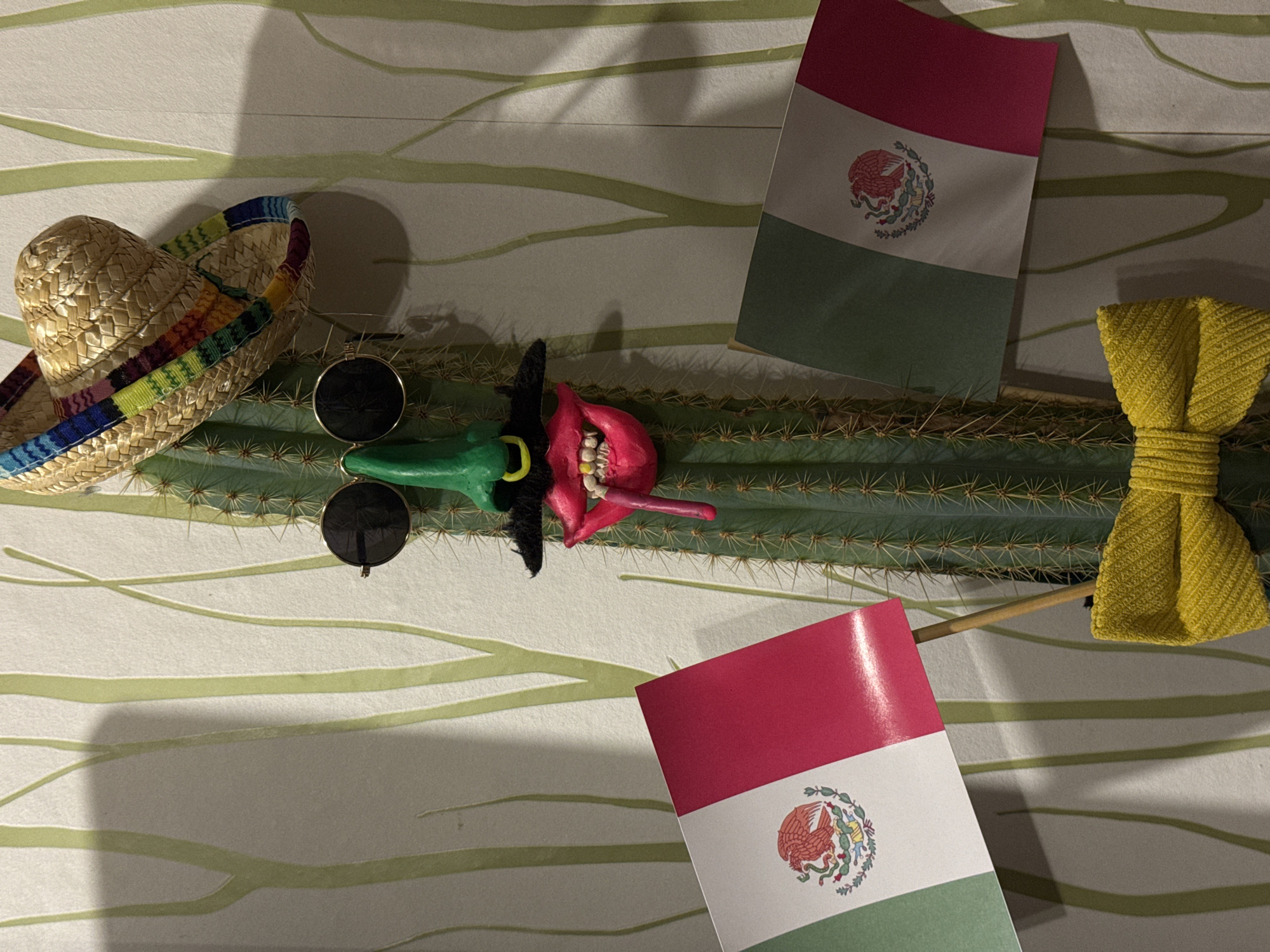 The image displays a flat-lay arrangement of objects with a Mexican theme. A straw sombrero with a multicolored band lies to the left. A green cactus with visible spines stretches across the center of the image. On the cactus are sunglasses, a red and green painted figure, and two Mexican flags on sticks. A yellow woven bow tie is on the right. The objects are placed on a patterned surface with horizontal lines. The location is not identifiable as Ketoshi, Russia, and no people, actions, or interactions are depicted.