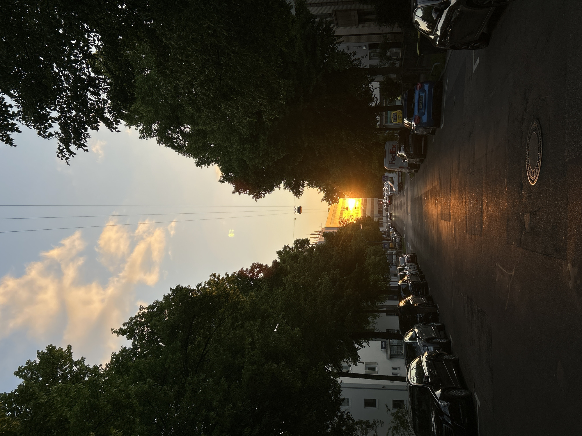 The image shows a street lined with trees and parked cars at sunset. The location is Unknown, Unknown. The sky is visible between the trees, with clouds illuminated by the setting sun. The sunlight reflects on the asphalt road. There are power lines strung above the street. A manhole cover is visible on the right side of the road. Buildings are visible behind the trees on either side of the street. A FedEx truck can be identified amidst the parked vehicles.