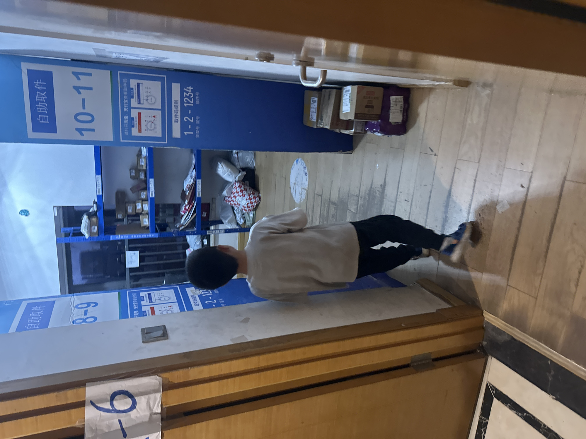 In a narrow hallway with wooden floors, a young boy wearing a grey long-sleeved shirt and dark pants walks away from the camera. To his right, a tall blue cabinet with white lettering is positioned against a wall. The cabinet has sections labeled "自助取件" (Self-service pickup) with numbers "8-9" and "10-11" in large blue font. Below these, there are smaller instructions and a code "1-2-1234". Stacked on the floor to the right of the cabinet are several cardboard boxes, some bound with purple packaging. To the boy's left, another white panel with a large blue "6" is partially visible. Shelving units filled with packages are visible in the background. The scene appears to be an indoor location, possibly a distribution or pickup point in the Chongqing High-tech Zone, China, given the visible Chinese text.
