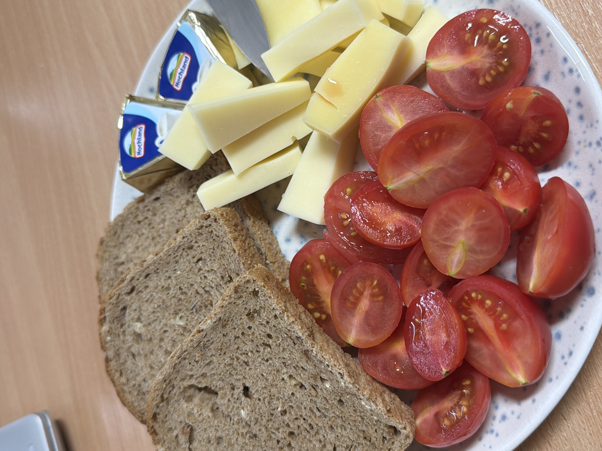 An overhead shot displays a simple meal or snack arranged on a white plate with grey speckles, set against a warm, light brown wooden surface. The plate is positioned on the right side of the frame, showing a variety of food items.
In the upper left quadrant of the plate, there are several irregular cubes and slices of light yellow cheese. A silver knife tip is visible, suggesting the cheese has been freshly cut or is being served. Next to the cheese, two small, rectangular foil packets of cheese spread are visible. These packets have a distinctive blue and gold design, with the brand name "Hochland" clearly printed on them, along with a logo featuring a mountain silhouette.
To the right, a generous portion of halved cherry tomatoes is artfully arranged. The tomatoes are bright red with glistening, juicy interiors, showing their seeds and pulp. Many of them are cut lengthwise, while some appear to be cross-sections.
On the left side of the plate, partially extending onto the wooden surface, are three slices of dark brown bread, possibly whole wheat or rye bread, showing a dense texture.
The scene depicts a prepared snack or light meal, possibly breakfast, ready for consumption. The location context provided is Sofia, Bulgaria. No people are visible, and the time of day is not explicitly indicated by lighting.