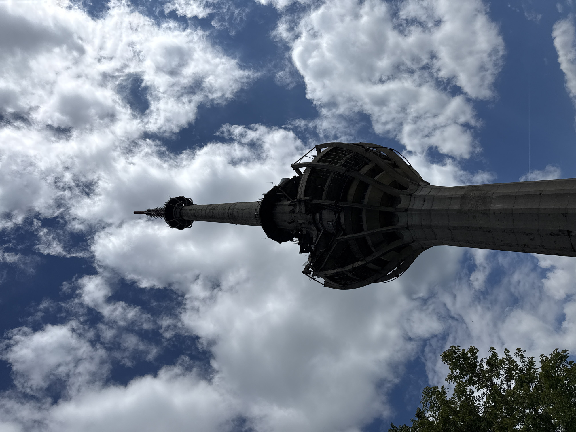 An upward shot of the Avala Tower in Sremska Kamenica, Serbia. The tower's concrete support structure is visible, ascending toward its distinctive latticed, rounded section. The camera angle emphasizes the tower's height. The background is a sky with cumulus clouds and patches of blue sky. Trees with green leaves are visible in the lower right corner.