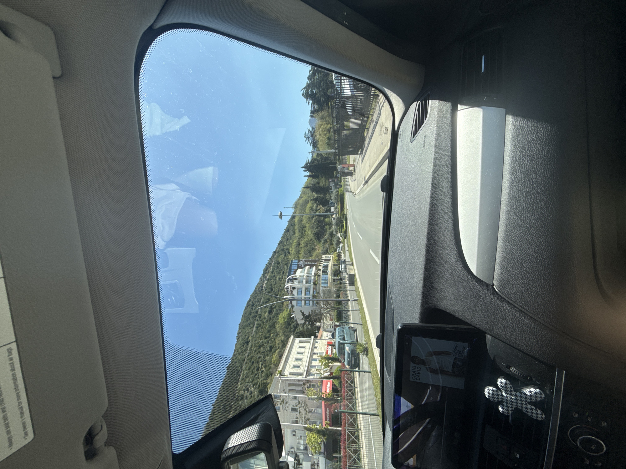 The image, taken from inside a car, shows an upward-sloping street in Unknown, Unknown, on a sunny day. The view is through the windshield, where the sky is blue, and a mountain covered in greenery dominates the landscape. A two-lane road runs along the base of the mountain, lined with several buildings of varying sizes and architectural styles.
Inside the car, the dashboard is visible, with a screen displaying an image and a houndstooth-patterned air freshener. The sun visor is down on the left side of the windshield. The scene suggests the car is traveling uphill on a clear day in a residential area near the mountainside.