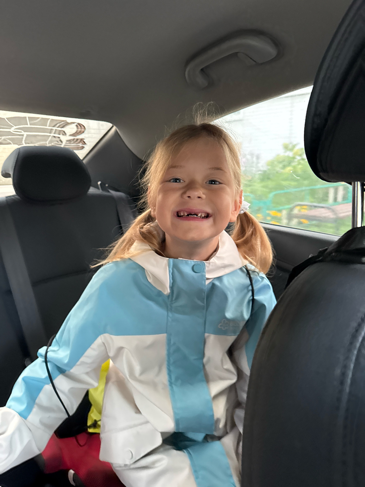 A young girl with blonde pigtails is sitting in the back seat of a car. She is wearing a light blue and white jacket. The girl is smiling and looking at the camera. The interior of the car is visible, including the back seats and the roof. Outside the car, trees and a building are visible through the window. The image appears to have been taken in Chaikovsky, Russia.