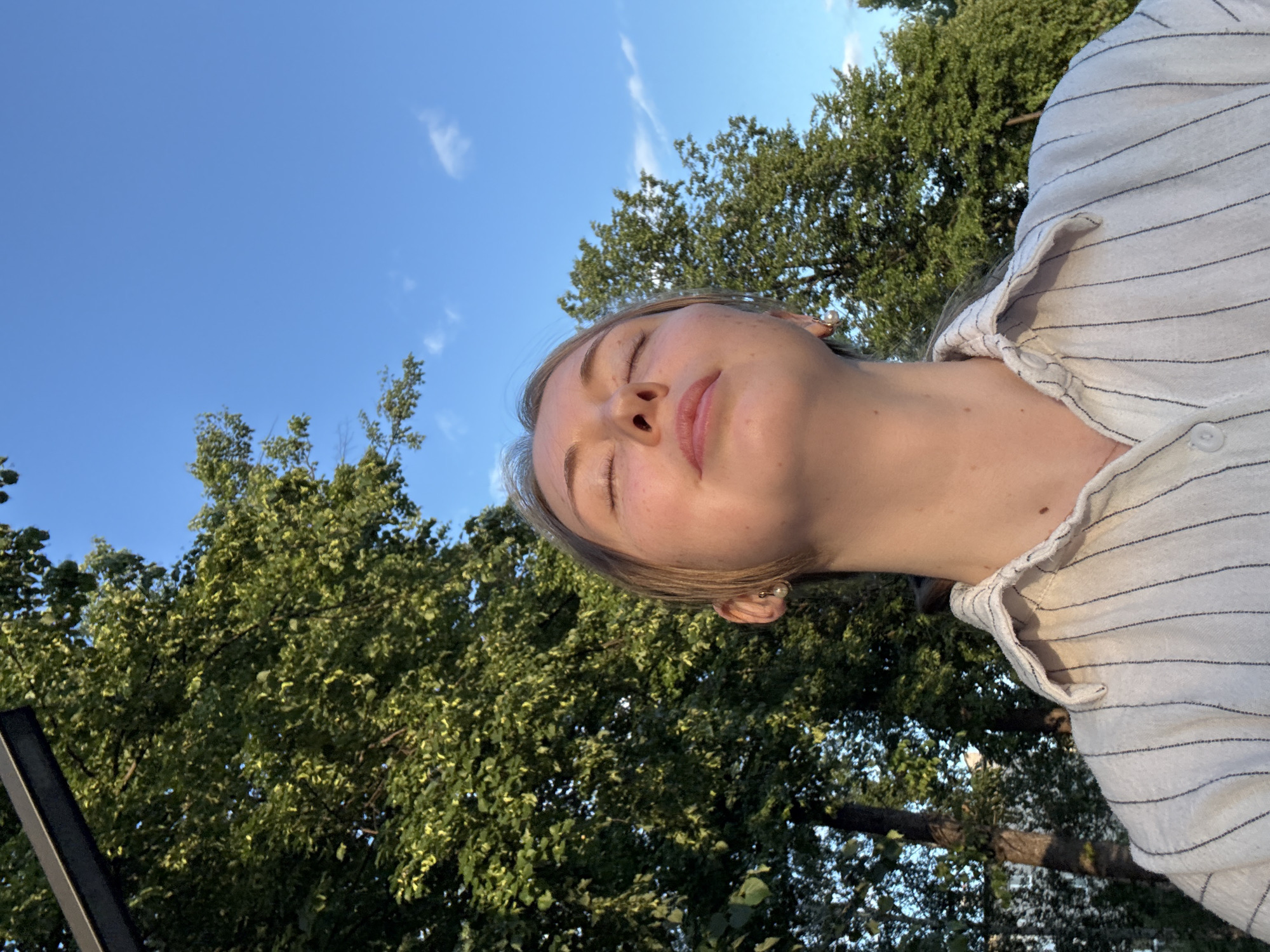 A young woman with blonde hair is captured in a close-up, low-angle outdoor shot, her eyes closed as she tilts her head back. She is wearing a white collared shirt with thin dark vertical stripes and a small pearl earring in her visible ear. The background features lush green trees with sunlight filtering through the leaves, and a clear blue sky with a few wispy clouds. The scene appears to be in a park or natural area within a city, with a dark, rectilinear structure possibly a lamppost or architectural element visible in the upper left corner. The lighting suggests it is late afternoon or early evening.