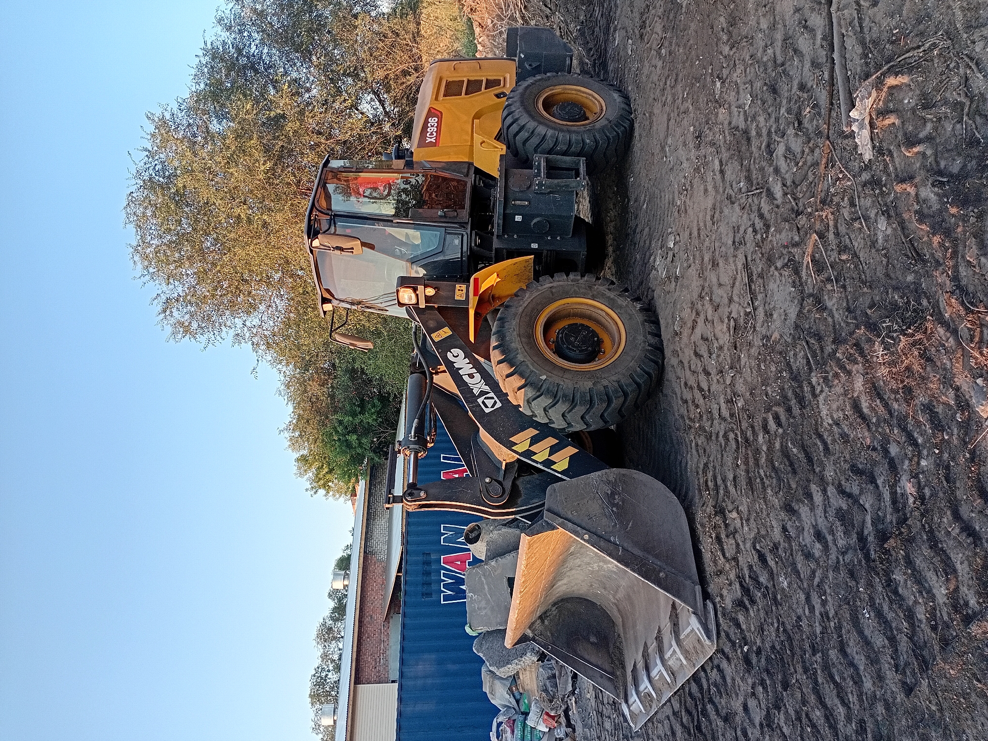 An XCMG XC936 wheel loader is parked on a dirt surface. The loader is yellow and black. It has a large bucket in the front, which is partially filled with debris. Behind the loader, there is a blue shipping container with the word "WAN" visible. There are also piles of debris and some trees in the background. The sky is clear and blue. The scene appears to be in an outdoor industrial or construction area.