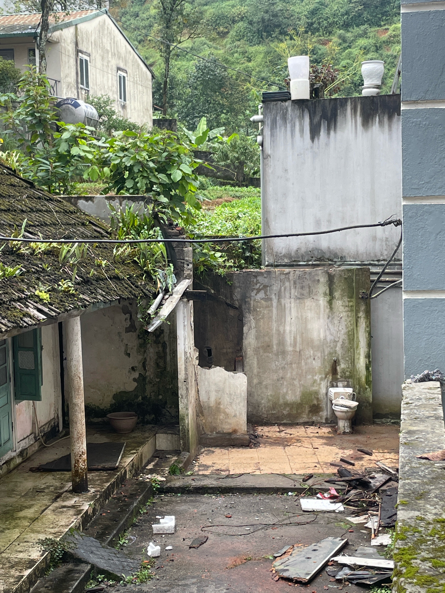 The image depicts a cluttered outdoor area in Sa Pa Ward, Vietnam. In the foreground, a damp, uneven ground is strewn with debris including pieces of wood, tiles, and what appears to be some white packaging. A small, weathered white object, possibly a concrete block, sits on the ground. A section of dark, angled material covered in green moss is also present.
To the left, a covered walkway or porch area is visible, with a weathered concrete pillar and a partially open green shuttered window. A brown, basin-like object rests on the floor. The roof of this structure is moss-covered and has vegetation growing on it, with a damaged metal gutter.
In the mid-ground, a light-colored, weathered concrete wall stands, showing signs of staining and discoloration. Against this wall, two toilets are placed, one of which appears functional with a lid. The area around the toilets is also uneven and somewhat dirty. Further back, a larger, light grey concrete structure rises, with some vertical pipes visible on its left side and what looks like a water tank on top. Two white objects, possibly planters or decorative items, are on the roof of this structure.
In the background, a multi-story building with a pale facade and several rectangular windows is partially visible. Lush green vegetation covers the sloping hillsides behind the structures. A water tank with the word "Suki" is visible on the side of the building in the background, surrounded by greenery. The overall impression is one of an aged, somewhat neglected environment, possibly a residential area or a service area, with evidence of decay and debris.
