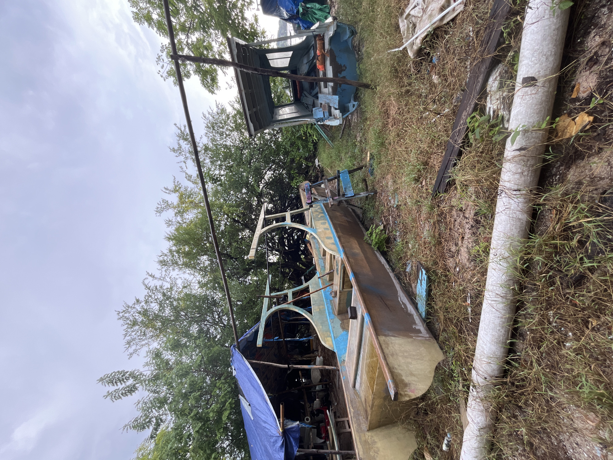 The vertically oriented image captures an outdoor, somewhat disheveled scene, likely a makeshift boatyard or storage area in an unknown part of Indonesia. The setting features abundant green foliage from trees dominating the left and right sides, and overgrown grass and weeds covering the uneven ground.
In the foreground and mid-ground, a prominent light brown or beige boat, appearing to be under construction or undergoing repairs, rests on the ground. It has a distinctive, arching frame structure on its deck, accented with traces of blue paint. To the left, a large blue tarp partially covers an area with various objects and supports, possibly sheltering another small boat or equipment. Further in the background, to the right, a smaller, older blue boat or boat-like structure is visible, partially obscured by vegetation and other materials.
The ground is littered with miscellaneous items, including a long, grey pipe running horizontally across the lower part of the frame, wooden planks, and scattered debris. Metal poles, possibly part of a support structure or scaffolding, are also visible. The sky above is uniformly overcast, a light grey, suggesting a daytime scene under cloudy weather conditions. No people are present, and no active event is taking place, but the environment strongly suggests activities related to boat building, repair, or long-term storage. No visible text is present in the image.
