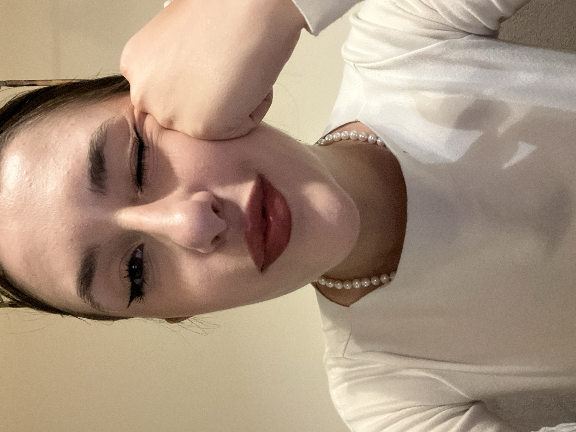 A young woman with dark hair and eyebrows is shown in a close-up, tilted shot. She is wearing a white top and a pearl necklace. Her left eye is closed, and her right eye is open with visible eyelashes and eyeliner. Her face is resting on her left hand, and her lips are glossy. The background is a plain, light beige color. There is no indication of the location being Sofia, Bulgaria.