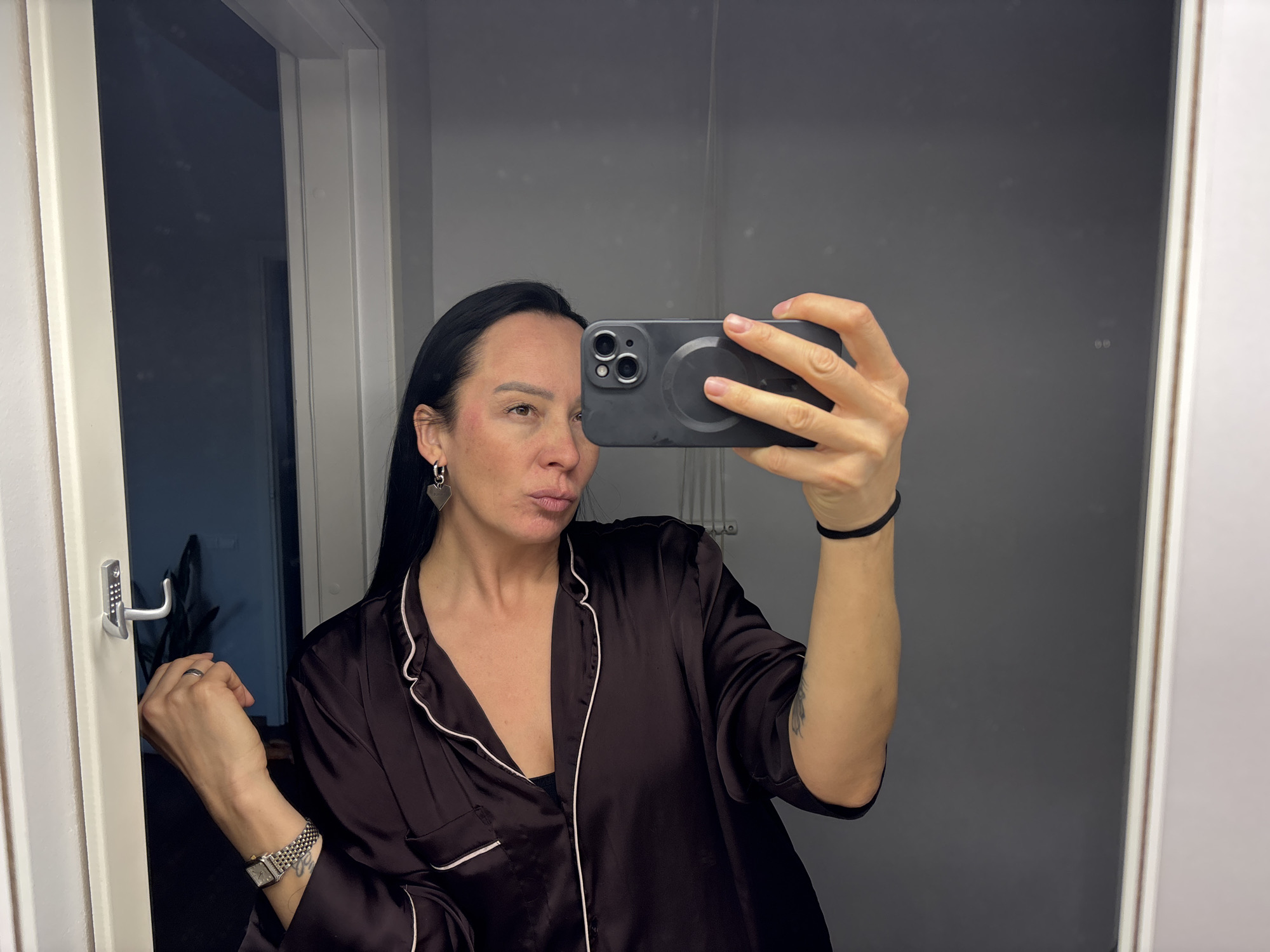 A woman with long black hair and a dark satin pajama top is taking a selfie in a bathroom. She is holding a black smartphone with a circular magnetic attachment on the back. She has a silver watch on her left wrist and a black hair tie on her right wrist. She is wearing heart-shaped earrings. The bathroom has a gray wall and a white-framed door. The exact location in the city of Chaikovsky, Russia is not identifiable.
