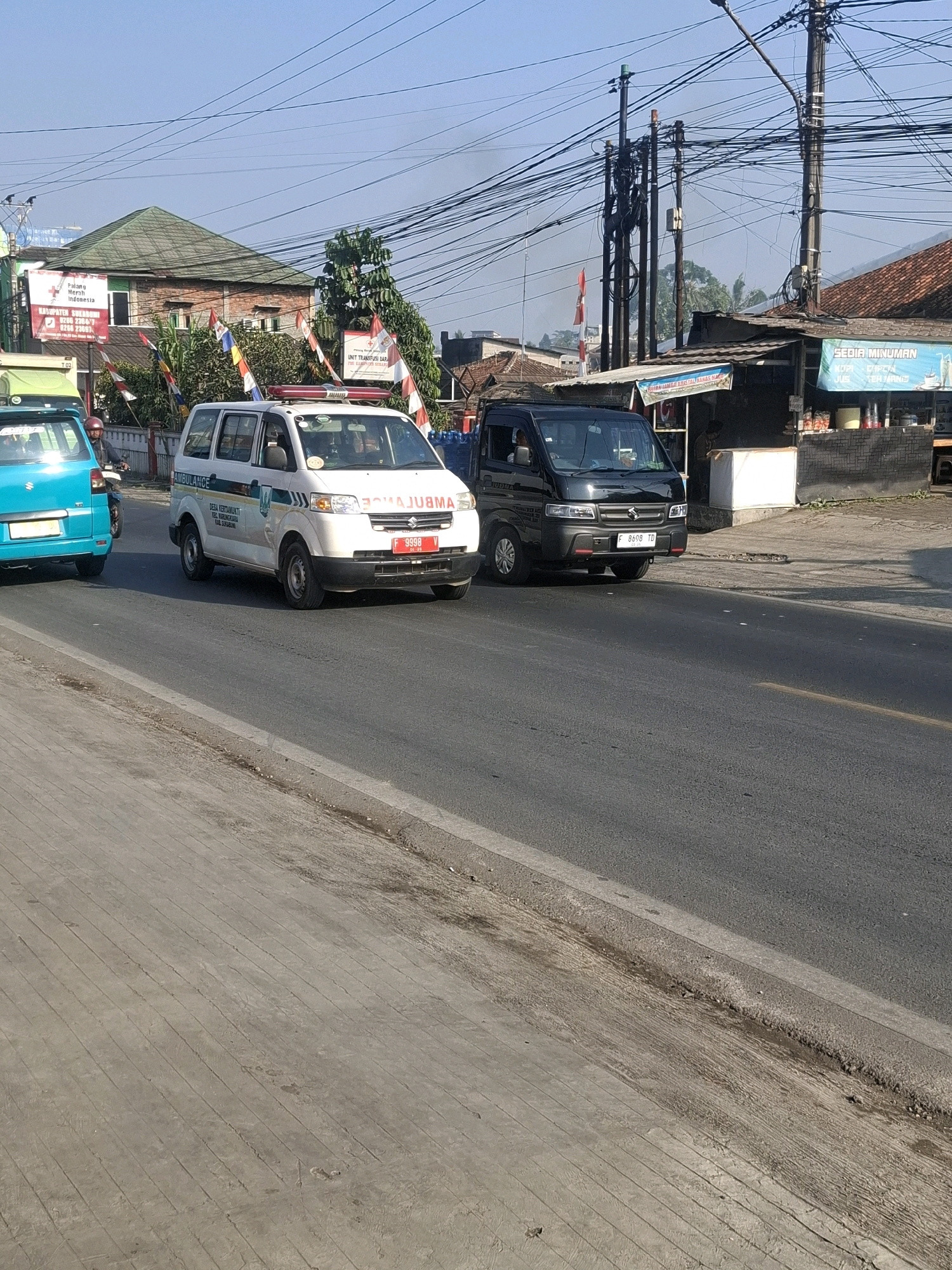 In Selajambe, Indonesia, a bright blue car is parked on the left side of the street. To its right, a white ambulance with red and blue lights on its roof is driving, its side facing the camera. The ambulance has text on its side that reads "AMBULANCE" and "DESA KERTAMUKTI KEL. BUKANGKARA KAB. SUKABUMI". Behind the ambulance, a black utility vehicle is also driving. The street is paved with asphalt, and a concrete sidewalk runs along the left side of the road. Buildings line the right side of the street, including a store with a sign that says "SEDIA MINUMAN". Overhead, a dense network of power lines crisscrosses the sky. The sky is clear and blue.