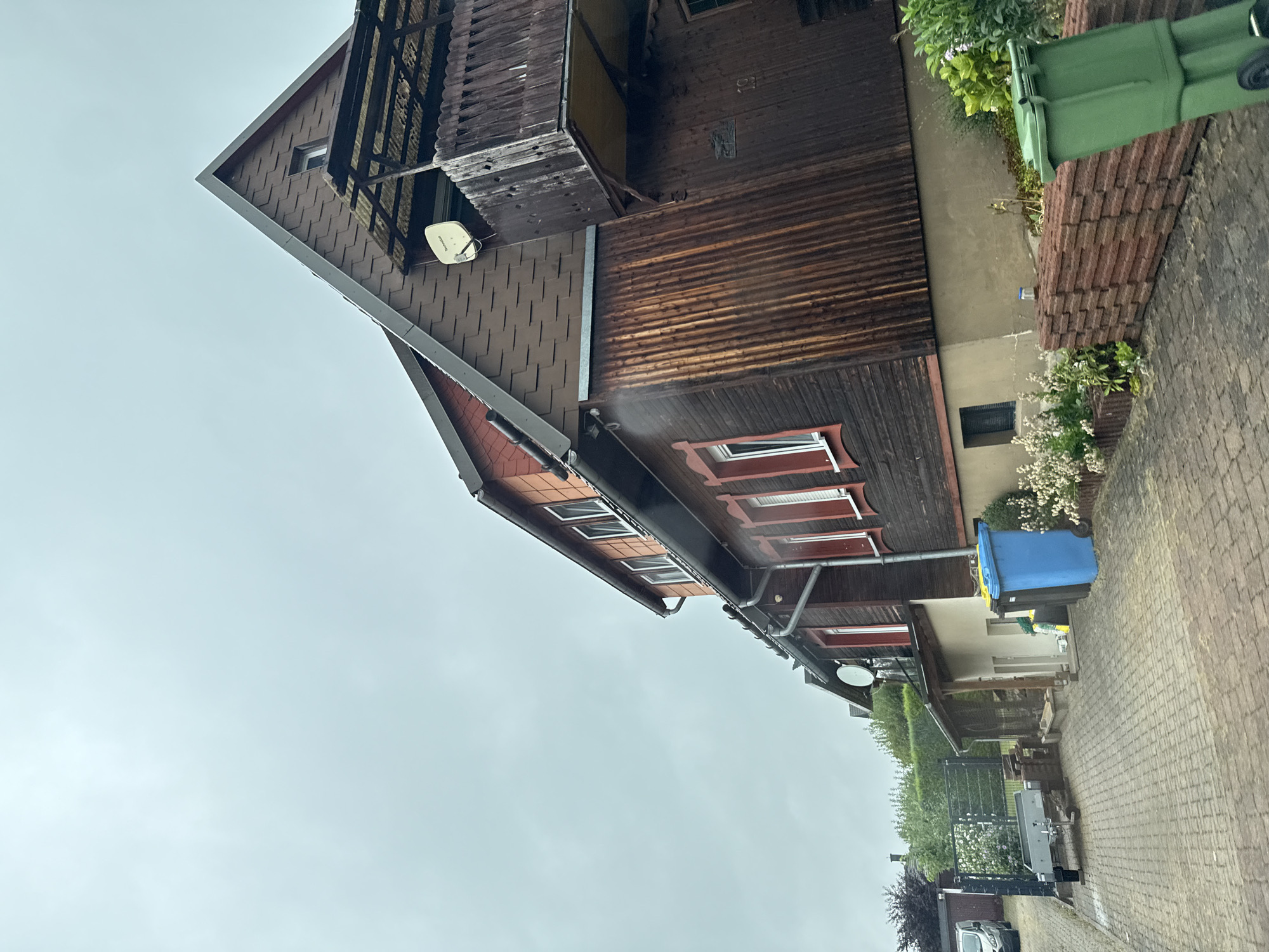 The image depicts the side and rear exterior of a multi-story building with dark wooden siding and a brown shingled roof. The building has several windows with red trim. A balcony with a wooden railing is attached to the upper level. A satellite dish is mounted on the side of the house. In the foreground, a paved driveway leads past two garbage bins, one blue and one green. A small trailer is parked on the left side of the driveway. Landscaping includes bushes and flowering plants. The sky is overcast. The scene is located in the town of Bad Sachsa, Germany.