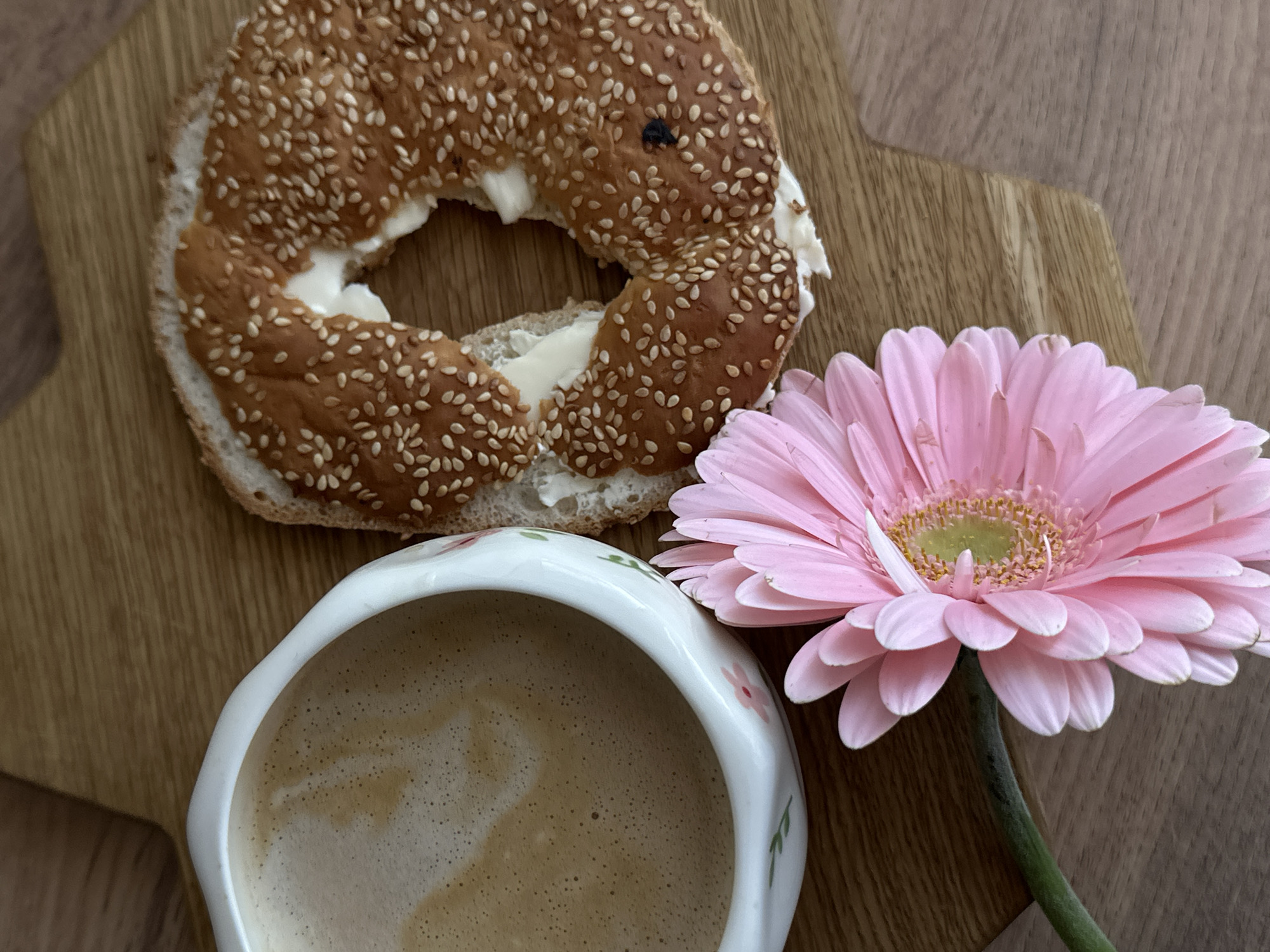 On a wooden surface, possibly a tabletop, lies a half-eaten sesame bagel filled with a white spread, next to a cup of coffee or cappuccino, and a pink gerbera daisy. The scene is indoors and could be in a cafe or home setting, but there are no identifying details to establish a specific location in Unknown, Unknown.