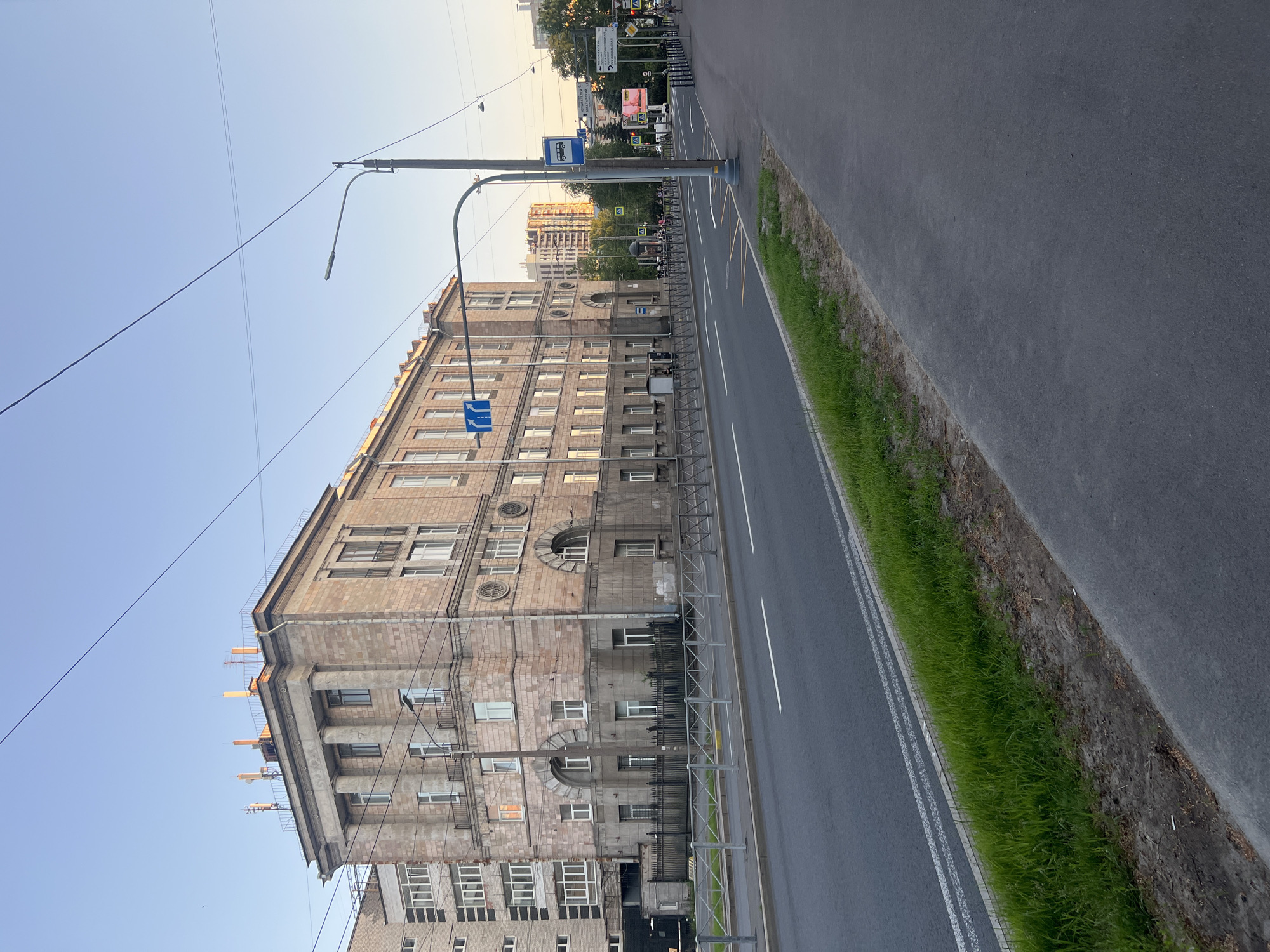 This image depicts a wide, multi-lane street in Saint Petersburg, Russia, observed during late afternoon or early evening, indicated by the soft golden light illuminating the facade of the primary building and the clear, pale blue sky.
On the left, a prominent multi-story building (approximately five stories) exhibits Neoclassical or Stalinist Empire architecture. Its facade is constructed from large, rusticated, pale reddish-brown stone blocks. The upper floors feature a central portico with at least four large, smooth columns supporting an ornate entablature and cornice. The ground floor displays several large arched windows, while the upper floors have rectangular windows, some with small, dark-colored Juliet balconies. The roofline includes a decorative railing, and several telecommunication antennas are visible on the rooftop. Adjacent to this building's left side, a modern, lighter-colored residential building with numerous rectangular windows is partially visible. A dark metal fence separates the classical building's immediate frontage from the road.
The street itself is paved with dark asphalt, featuring two visible lanes marked by white lines, including a double solid line and a single solid line. A narrow grass median with some soil is positioned between the road and a paved sidewalk. Metal pedestrian barriers with a diamond pattern line the edge of the road, separating it from the grass and sidewalk.
On the right side of the street, a tall dark-colored pole supports a modern streetlamp that extends over the roadway. A blue sign with a white bus icon marks a bus stop. Below it, a white rectangular directional sign with black Cyrillic text lists several street names: "ИНСТИТУТ АТЕЛЬЕ 4," "ул. БЕЛОРУССКАЯ," "УЛ. СМОЛЕНСКАЯ," "ул. БРЯНСКАЯ," "УЛ. ЛИГОВСКАЯ," "ул. БОРОВАЯ," and "УЛ. РУЗОВСКАЯ." Another blue traffic sign with white arrows indicating "straight" and "right" is visible on the left side of the street, above the fence.
In the distance along the street, trees, additional buildings (including taller, more contemporary structures), and several pedestrians are visible. Multiple overhead utility wires crisscross the sky. The overall scene conveys an active but not overly congested urban environment.
