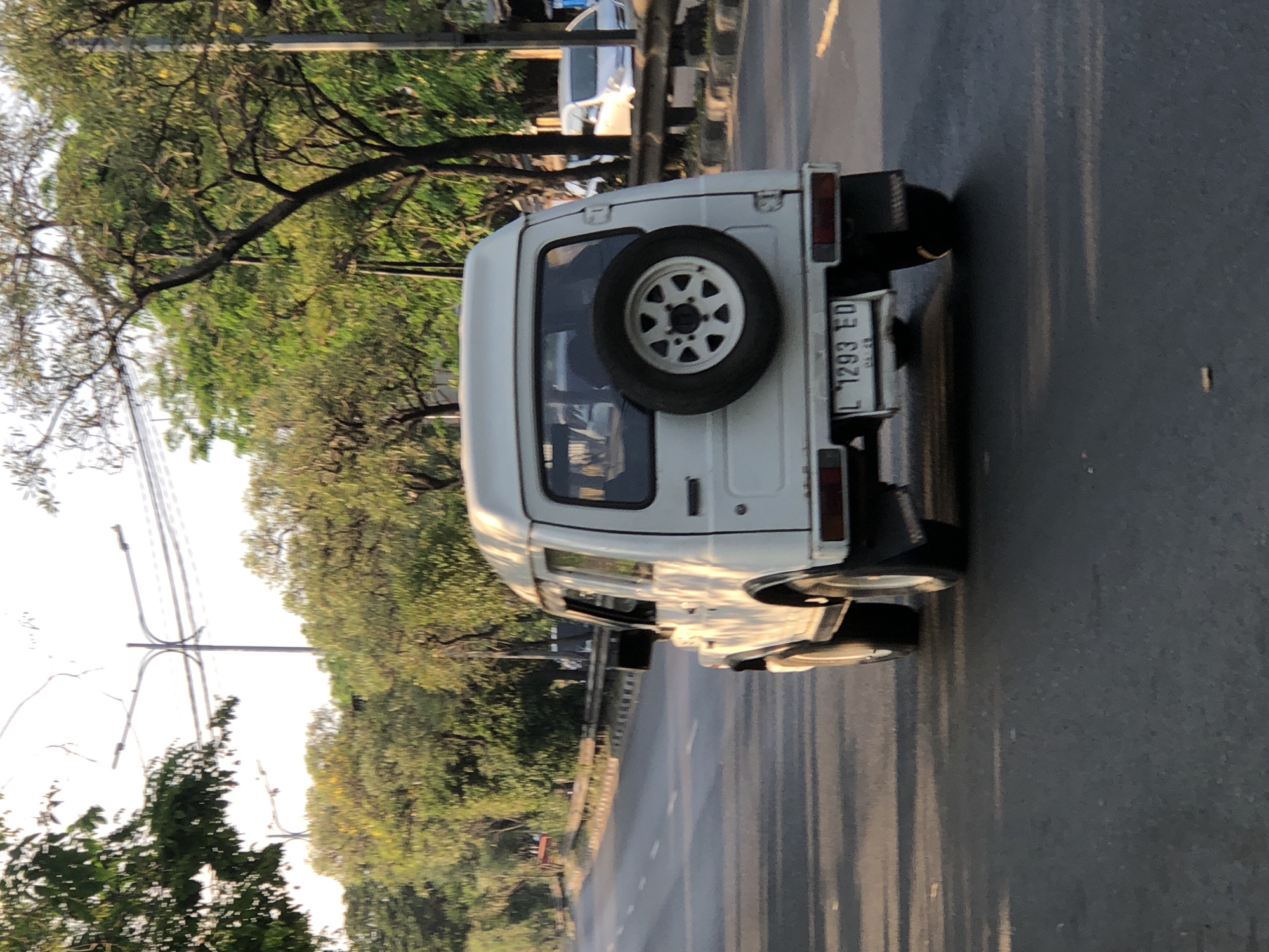 A white four-door SUV is driving on a paved road in Kludan, Indonesia, as suggested by the license plate reading "L 1293 ED". The SUV has a spare tire mounted on the back. The sun is low in the sky, casting long shadows on the road and illuminating the trees lining the street. A guardrail is visible along the side of the road. In the background, other vehicles and streetlights can be seen. The environment appears to be a suburban or semi-rural area.