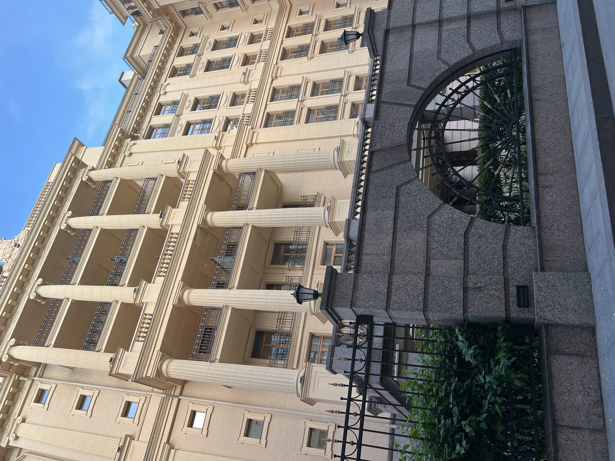 This image captures a low-angle view of a grand, pale yellow, multi-story building with classical architectural elements in Saint Petersburg, Russia. The building features numerous windows with dark frames, balconies with ornate railings supported by large, fluted columns, and decorative cornices. In the foreground, a dark metal fence with spiked finials runs horizontally, partially obscuring a lush green bush. A portion of the fence is set against a granite wall with an arched opening, revealing more of the building and foliage behind it. A black, ornate lantern stands on a granite pillar to the right of the fence. The sky above is a clear, light blue with a few wispy white clouds. The scene appears to be a street-level view of a residential or office building, characterized by its imposing and elegant design.