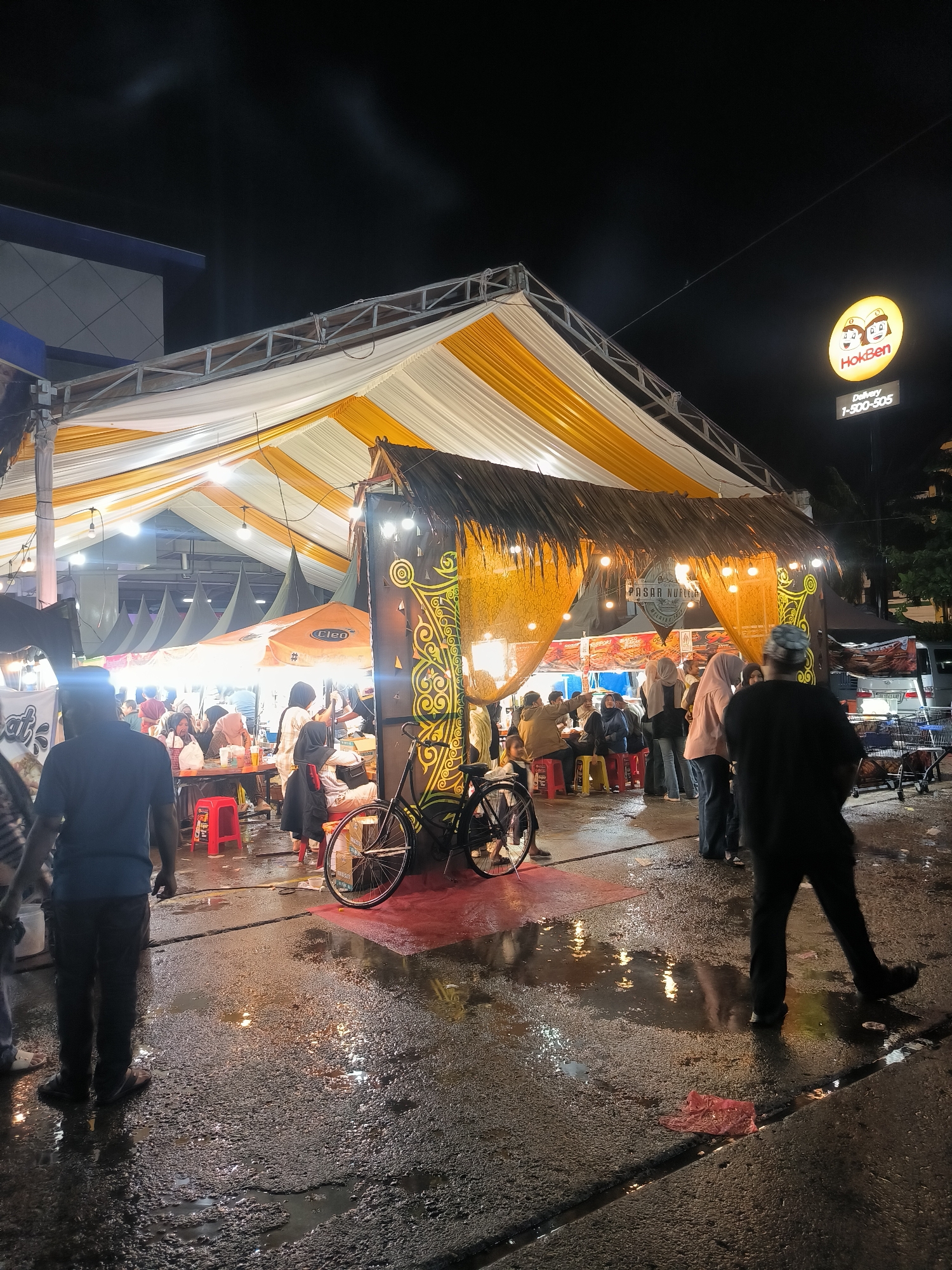 This nighttime image captures a busy outdoor market or food court in Lhokseumawe, Indonesia. A large, temporary tent structure with white and yellow fabric panels serves as the primary overhead cover for the area. Below this main tent, numerous individuals, predominantly adults, are engaged in various activities.
In the center-left, people are seated at small tables on red plastic stools, appearing to dine or socialize. Behind them, multiple dark, conical tent structures are visible, suggesting additional stalls. One prominent stall in the foreground features a thatched roof made of dried leaves and is adorned with decorative, traditional-looking motifs in yellow and black. This stall is lit by multiple bare-bulb string lights. A partially visible sign within this stall reads "PASAR NURAGA". An orange umbrella with the "CLEO" logo is also visible.
The ground is wet asphalt or concrete, marked with puddles and reflective surfaces that mirror the overhead lights, indicating recent rain or moisture. A red plastic bag lies discarded on the wet ground in the lower right.
Several individuals are standing and walking across the scene. In the immediate foreground-left, the back of a person in a blue shirt and dark pants is visible. To the right, another person in a dark shirt and pants walks away from the viewer. Many women in the scene wear hijabs.
A bicycle is parked near the thatched stall on the right. In the upper right background, an illuminated circular sign for "HokBen" is prominently displayed, with "Delivery 1-500-505" written below it. Further in the background, a white van and some shopping trolleys are visible near other market stalls. A modern building with blue and gray elements is visible on the far left. The overall scene is brightly lit by numerous artificial lights strung throughout the tent and stalls, creating a lively atmosphere.