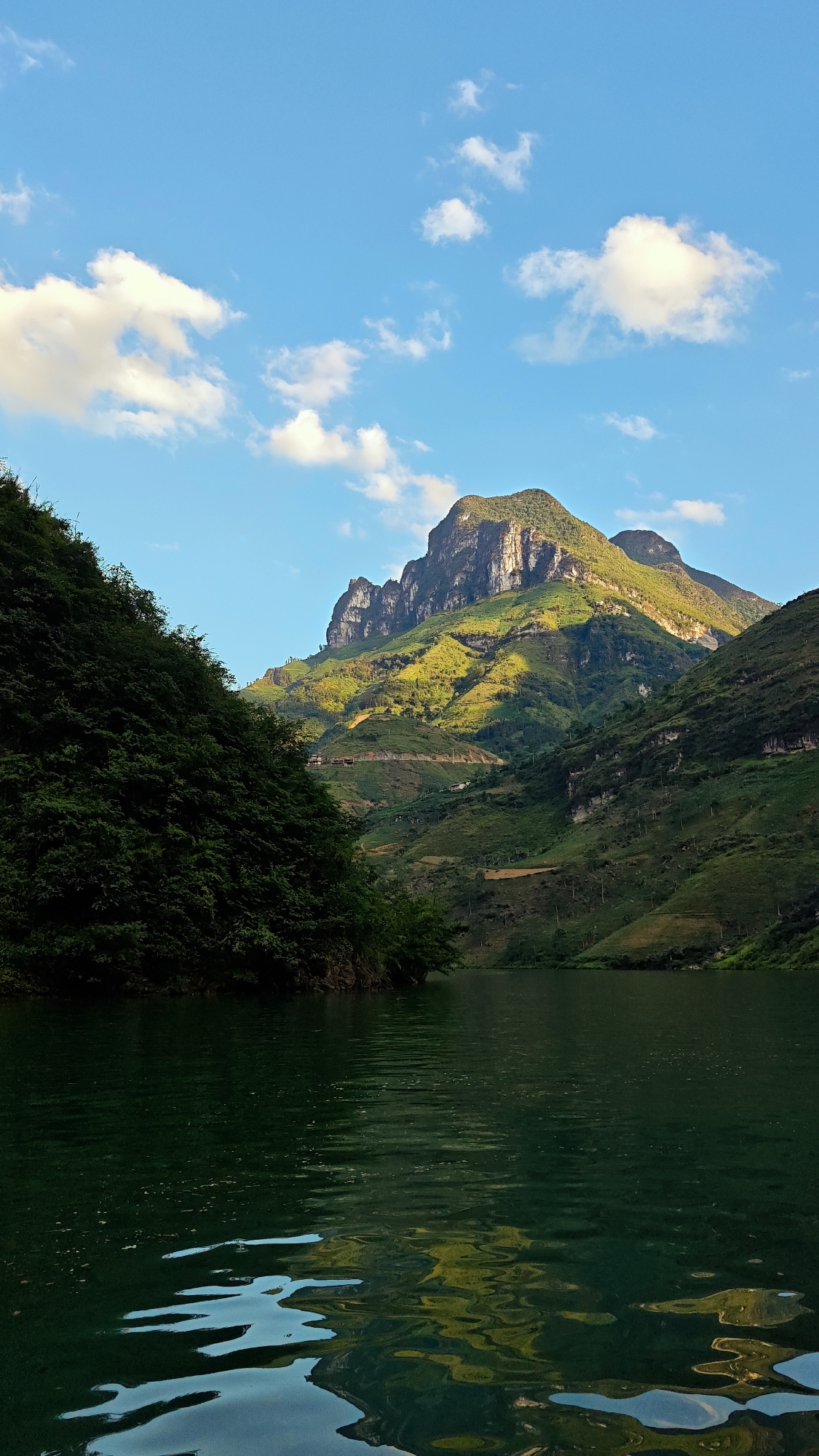 A striking landscape features a dark green lake in the foreground, reflecting the sky and surrounding terrain. On either side of the lake are steep, verdant hillsides densely covered with lush vegetation. In the background, a prominent mountain range rises majestically, partially illuminated by sunlight that highlights its rugged peaks and rocky cliffs. Narrow terraces and winding roads are visible along the mountainside, suggesting human cultivation and habitation. Above, a clear blue sky is punctuated by scattered white clouds. The scene appears to be in the Đồng Văn Commune, Vietnam, based on the distinctive landscape features.
