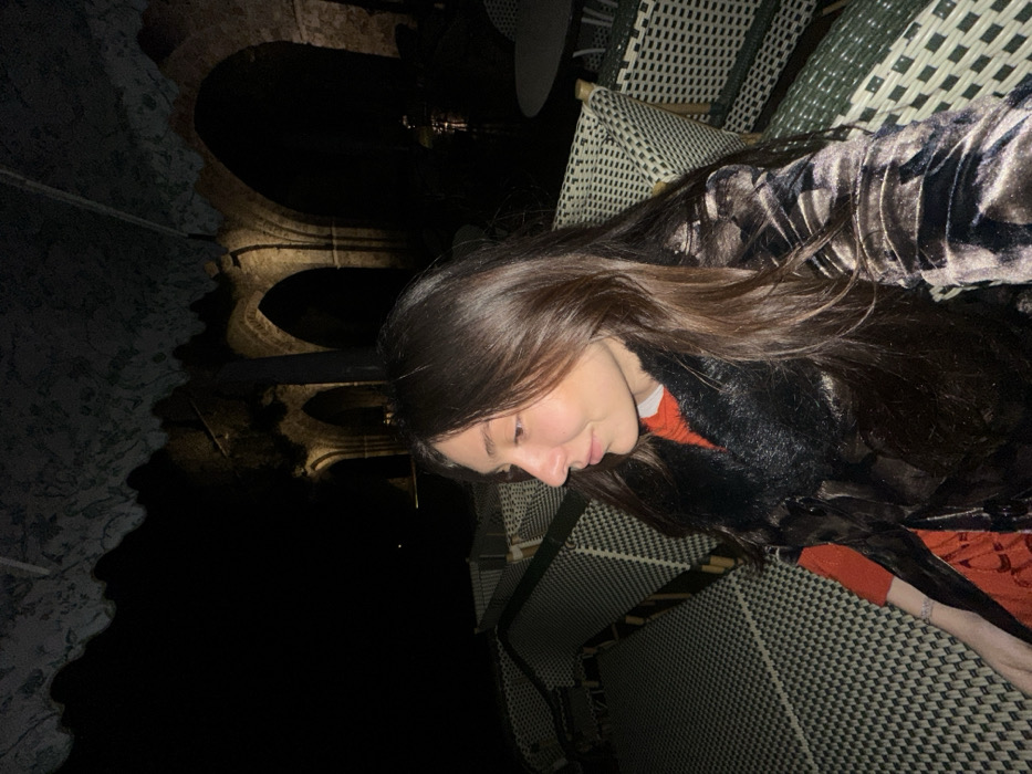 A woman with long brown hair is sitting outside on a patterned chair, wearing a dark velvet shirt over an orange top. She's looking downwards, seemingly lost in thought. In the background, the reflection of an arched structure can be seen in a dark body of water, suggesting a night scene near a canal or river with historic architecture. The image has a slightly rotated perspective.