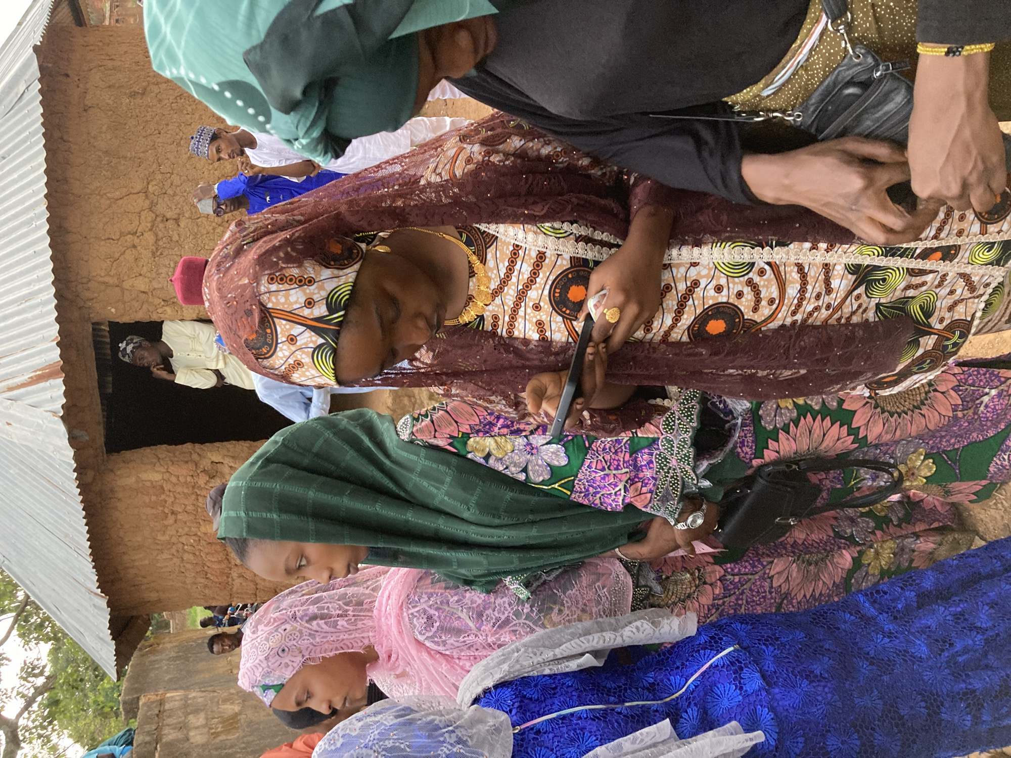 In Jos, Nigeria, a group of people, predominantly women adorned in colorful, patterned garments and headscarves, are gathered outdoors near a mud-brick structure with a corrugated metal roof. The focus is on several women in the foreground, dressed in vibrant fabrics with intricate designs. One woman, in the center, is wearing a maroon lace headscarf over a white and orange patterned dress and a gold necklace, and she is looking down at a mobile phone in her hands. To her left, a woman in a pink headscarf and a pink patterned dress is visible. To the right of the central figure, another woman wears a green headscarf and a dress with a floral pattern. A woman on the far left is in a blue, lace-like dress with a white headscarf. In the background, several men are seen, some in traditional attire, standing near the doorway of the building. The ground appears to be dirt.