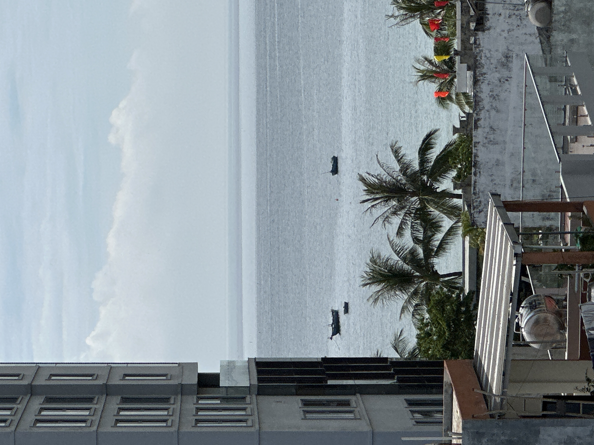 The image captures a view of the sea from a building, likely in Đà Nẵng, Vietnam. On the left, a tall, modern building with rows of windows dominates the foreground. To the right of this building, the sea stretches out to the horizon under a partly cloudy sky. Two small boats are visible on the water. In the middle ground, a line of palm trees and lush greenery separates the sea from a lower structure in the foreground. This foreground structure appears to be a rooftop with a concrete wall, a covered area with a striped awning, and a water tank. Colorful flags, red and yellow, are also visible on the rooftop in the mid-ground, adding a splash of color to the scene. The overall impression is one of a coastal urban environment.