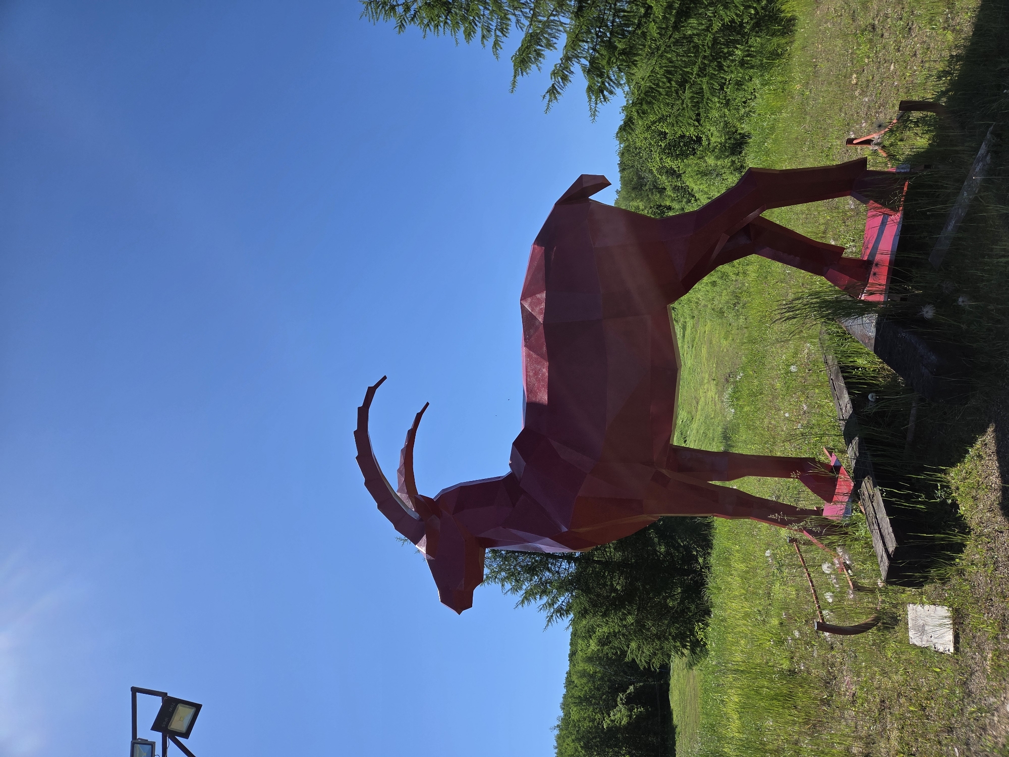The image displays a large, red, geometric sculpture of an ibex standing in a grassy field under a clear blue sky. The sculpture is metallic and appears to be made of faceted panels. The ibex is depicted in profile, facing left, with its head held high and its curved horns prominently featured. In the foreground, the ground is covered in green grass with some dandelion seeds. To the left of the sculpture, there is a metal object resembling a plow or a piece of farm equipment, and a wooden plank. To the right of the sculpture, a tall evergreen tree is visible. The background consists of a gentle, tree-covered slope. The sun is shining brightly, casting a slight glare in the upper left corner of the image. There are no people visible in the image. The location is not identifiable as Birobidzhan, Russia, based on the visual evidence.