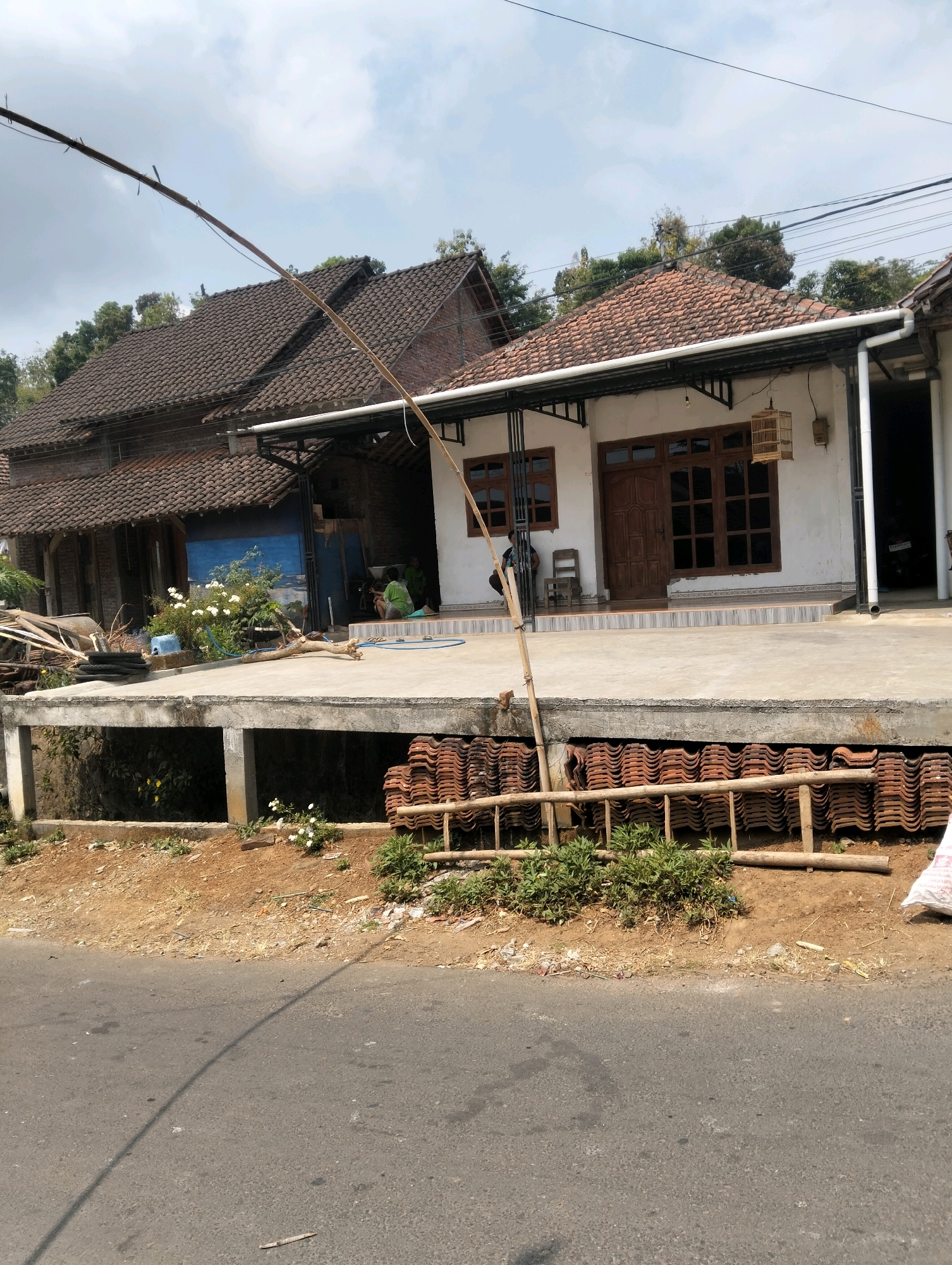 The image depicts a street view in Kanyoran, Indonesia, featuring a residential structure with an elevated concrete platform. The house has white walls and a tiled roof. A wooden birdcage hangs outside the house. In front of the house, a pile of terracotta roof tiles is stacked, supported by a wooden structure resembling a ladder or scaffolding. Vegetation, including small white flowers and green bushes, grows around the base of the platform and on the roadside. A paved road occupies the foreground, with a shadow cast across it. The scene suggests a rural or semi-rural setting, possibly indicating ongoing construction or renovation.