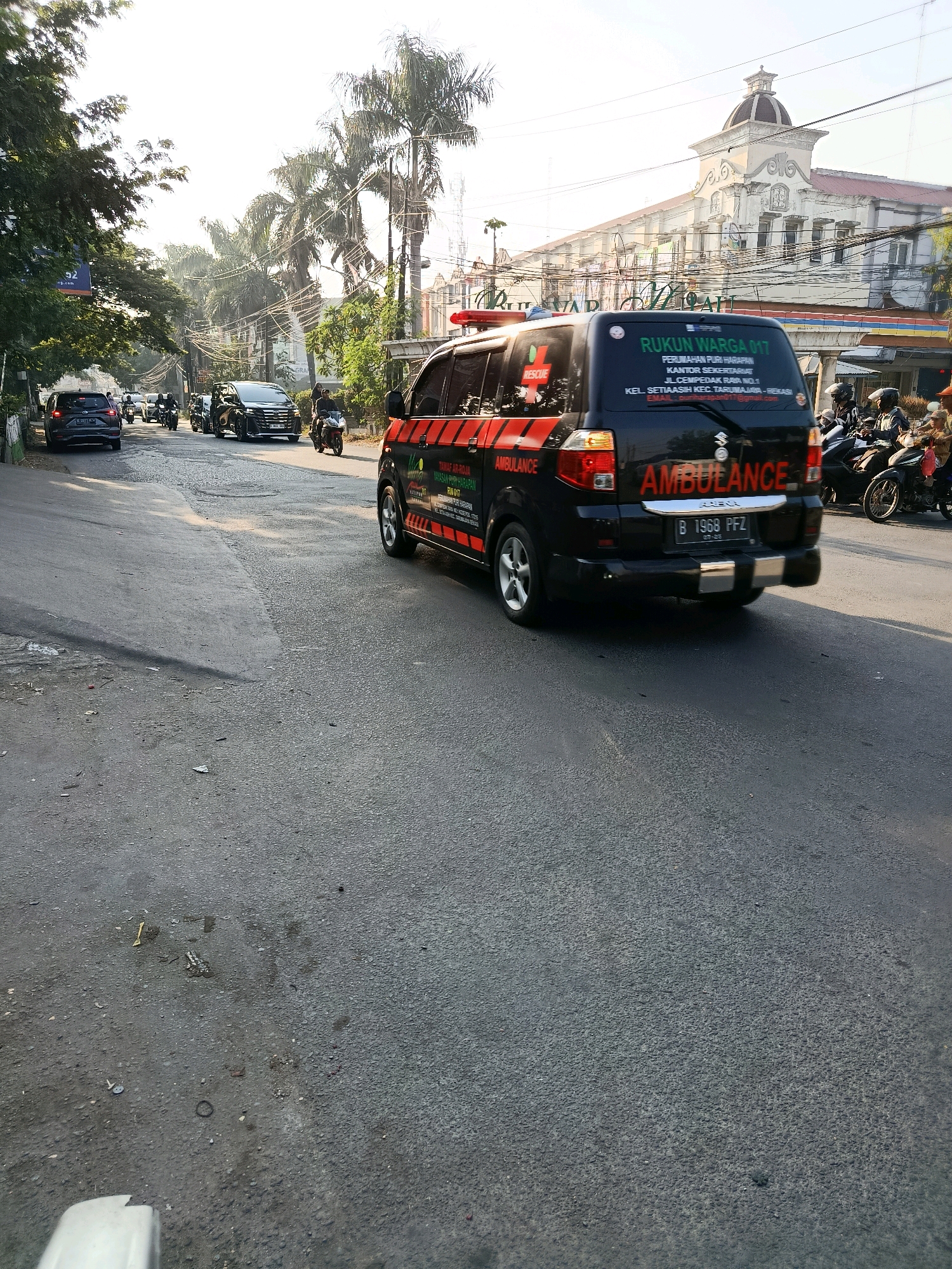 In Bekasi, Indonesia, on a road with a slightly uneven surface, a black Suzuki ambulance is captured in motion. The ambulance, branded with "AMBULANCE" in prominent red lettering and a red cross, displays "RUKUN WARGA 017" on its rear window, indicating its affiliation with a local neighborhood organization. Text below provides an address: "KEL. SETIAASIH KEC. TARUMAJAYA - BEKASI" and an email address. The ambulance has a license plate that reads "B 1968 PFZ." Several other vehicles, including cars and motorcycles, are present on the road, suggesting moderate traffic. Palm trees line the left side of the road, and a building with classical architectural elements, featuring a dome, stands in the background. The sky is bright, indicating daytime.