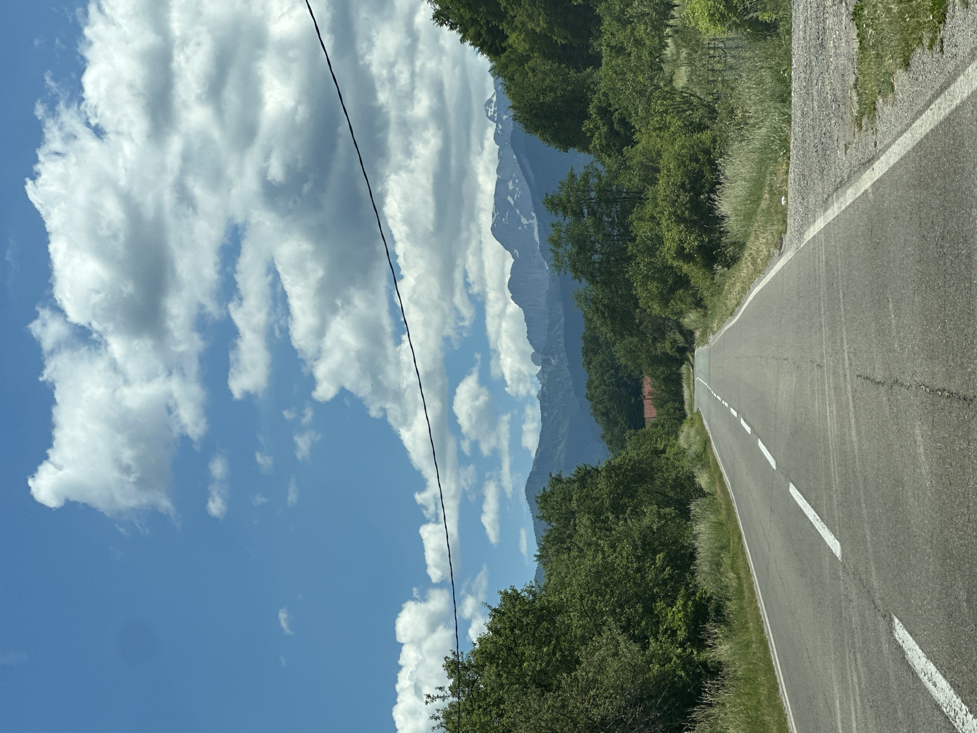 The image depicts a scenic road leading towards a mountainous landscape under a bright blue sky dotted with fluffy white clouds. The road is paved, with white dashed lines marking the lanes. Lush green trees line both sides of the road, with a denser forest in the background. In the distance, majestic mountains rise, their peaks dusted with snow. The overall impression is one of a serene and natural environment, likely in a rural or mountainous area. The location is identified as Murino, Montenegro.