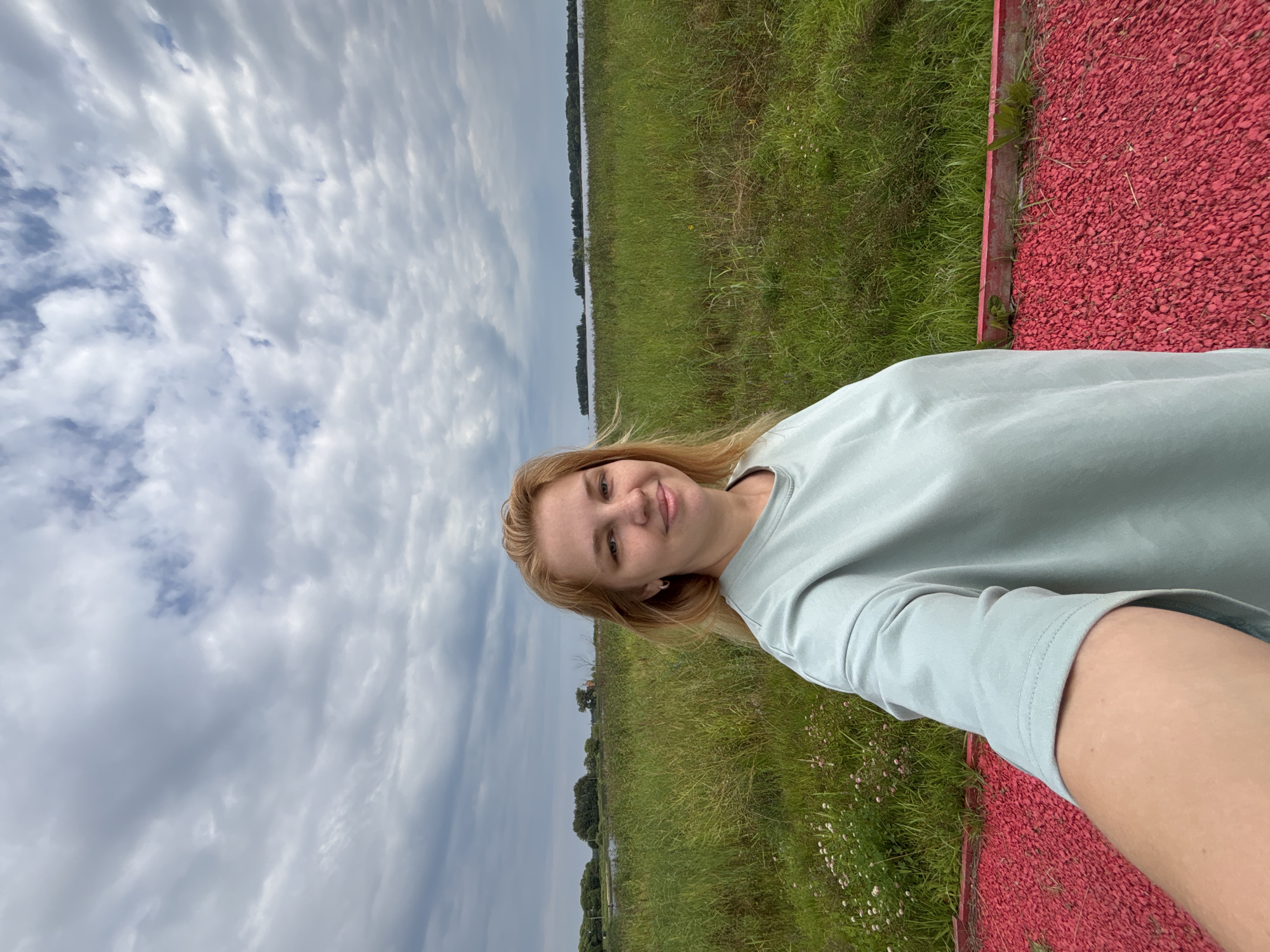A young woman with blonde hair and light skin is taking a selfie in an outdoor setting. She is smiling and looking at the camera. She is wearing a light blue long-sleeved shirt. In the background, there is a field of tall green grass and a body of water, likely a lake or river, with a treeline in the distance. The sky is cloudy. In the foreground, there is a red gravel path. The image appears to have been taken in the village of Breytovo, Russia, given the typical landscape and architecture often found in such rural Russian locations.
