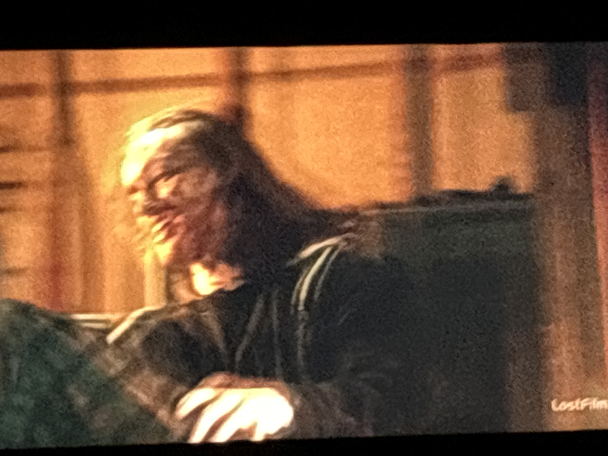 The image shows a disfigured orc-like figure with long dark hair, wearing a dark garment. The figure appears to be seated. The background is blurry, showing a warm-toned interior. The bottom right corner has the word "LostFilm". There is no indication of the location being Budva, Montenegro.