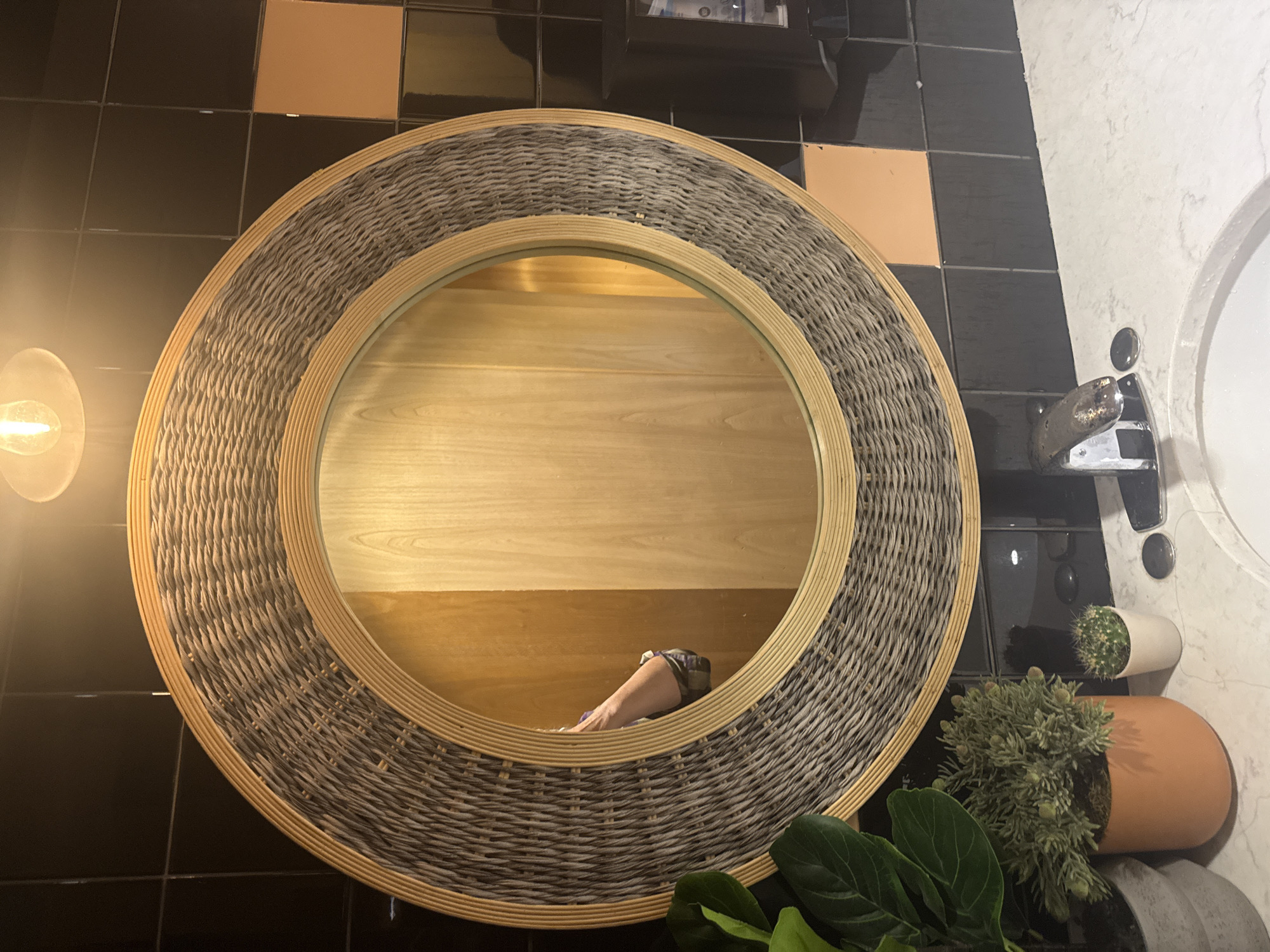 This image depicts a bathroom scene. A large, round mirror with a woven frame hangs on a tiled wall. The tiles are primarily black, with some accent tiles of a light brown or peach color. A light fixture is visible above the mirror. The mirror reflects a wooden door. Below the mirror, on a marble countertop, are two potted plants, one larger with green leaves and the other a small cactus. A faucet is visible on the countertop, positioned above a sink. The overall style of the bathroom appears to be modern with some natural elements. There is no identifiable information within the image that would indicate its specific location in Calgary, Canada.