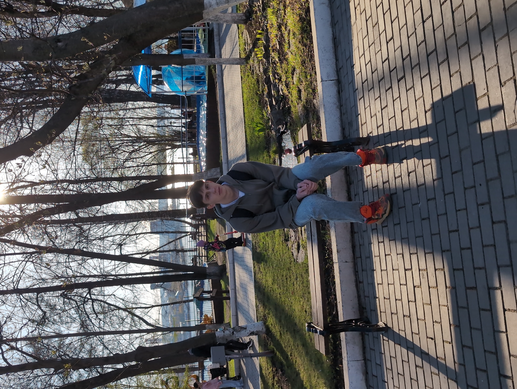 The image captures a young man sitting on a bench in Izhevsk, Russia, on a paved walkway. The man is wearing light blue jeans, an olive green jacket, and bright orange sneakers. He is looking directly at the camera. The bench is situated next to a strip of grass bordered by a concrete edge. Behind the bench, a park scene unfolds, with bare trees silhouetted against the sky. In the background, structures and possibly recreational features are visible. The sun is low in the sky, casting long shadows across the walkway. Other people can be seen in the distance, interacting within the park environment.