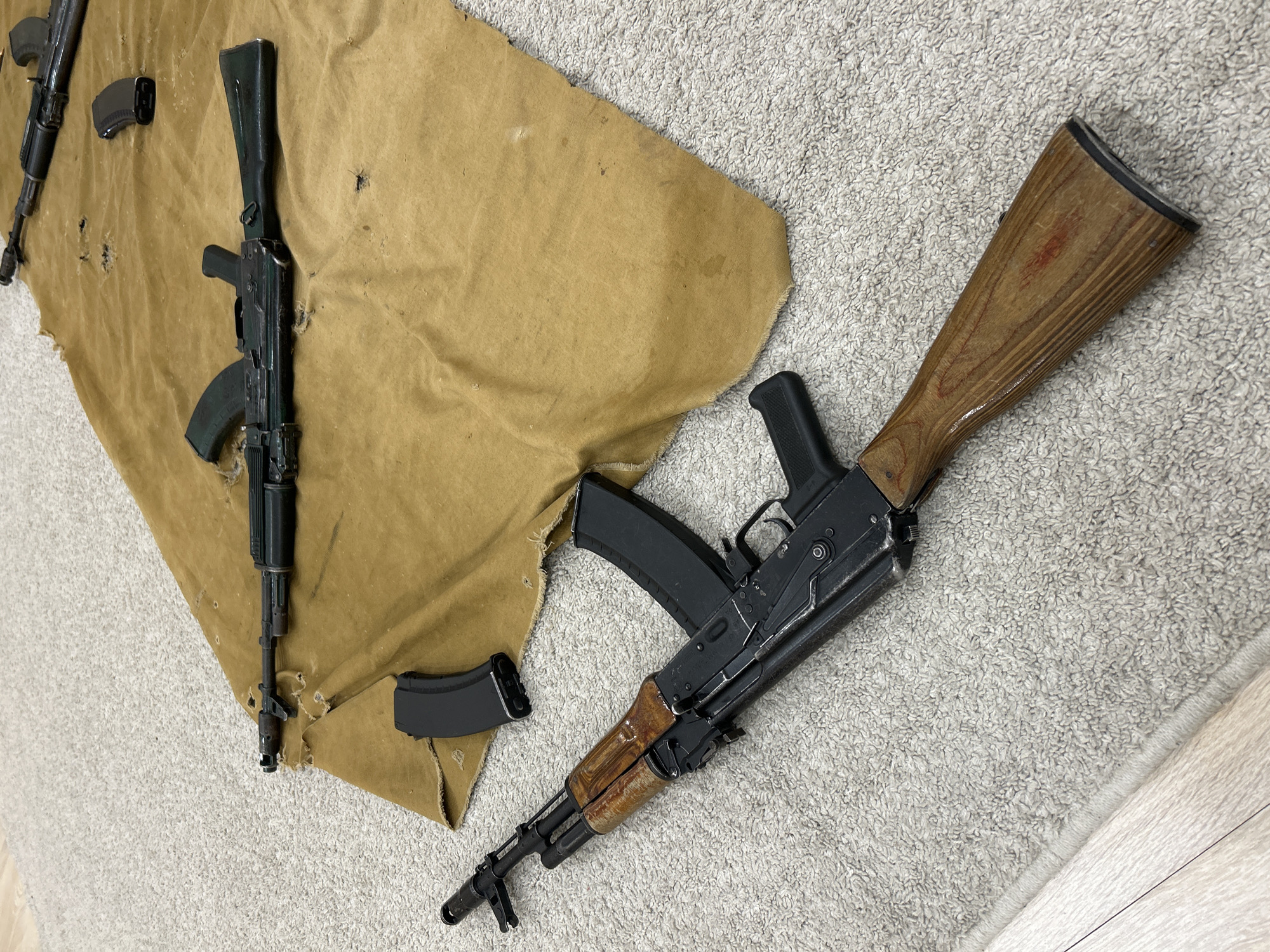 This media file presents an indoor scene featuring several firearms and accessories laid out on a combination of worn fabric and carpet. The primary subjects are three AK-style rifles and at least two detached magazines.
The most prominent rifle is positioned on the right side of the frame, showcasing a black metal receiver with a wooden buttstock and a wooden handguard. A black, curved magazine is inserted into its magwell. This rifle exhibits visible wear on both its wooden furniture and metal finish. A second rifle is partially visible on the left, also appearing to be an AK variant, with a darker (possibly polymer or painted wood) buttstock and handguard, and a magazine inserted. The barrel and gas block of a third rifle are barely discernible at the top left. Two additional black curved magazines are placed separately on the fabric.
The firearms rest on a large piece of light brown or tan fabric, resembling canvas or burlap, which displays considerable wear, including discoloration, stains, and multiple small holes. This fabric covers a portion of a light grey, shaggy-textured carpet. A small section of light-colored wooden flooring is visible in the bottom right corner, bordering the carpet.
No people are visible, and no specific activity is explicitly shown, but the arrangement implies the firearms are being stored, inspected, cleaned, or prepared. The items generally appear used but not heavily damaged. No visible text is present on any of the objects. While the location context is Izhevsk, Russia, the image itself does not contain specific visual cues to confirm this geographical location.