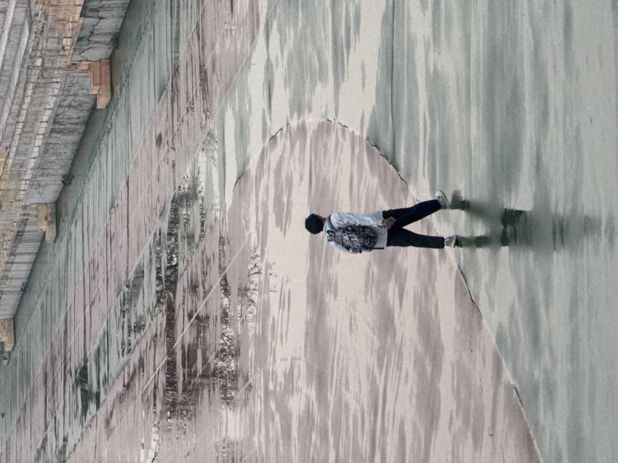 A person wearing a white jacket, dark pants, and white shoes is lying on the concrete surface next to the wall of a building. The scene is located in Zhongtun, China. The concrete has a pattern and is cracked. There is a reflection of trees and the sky on the concrete.