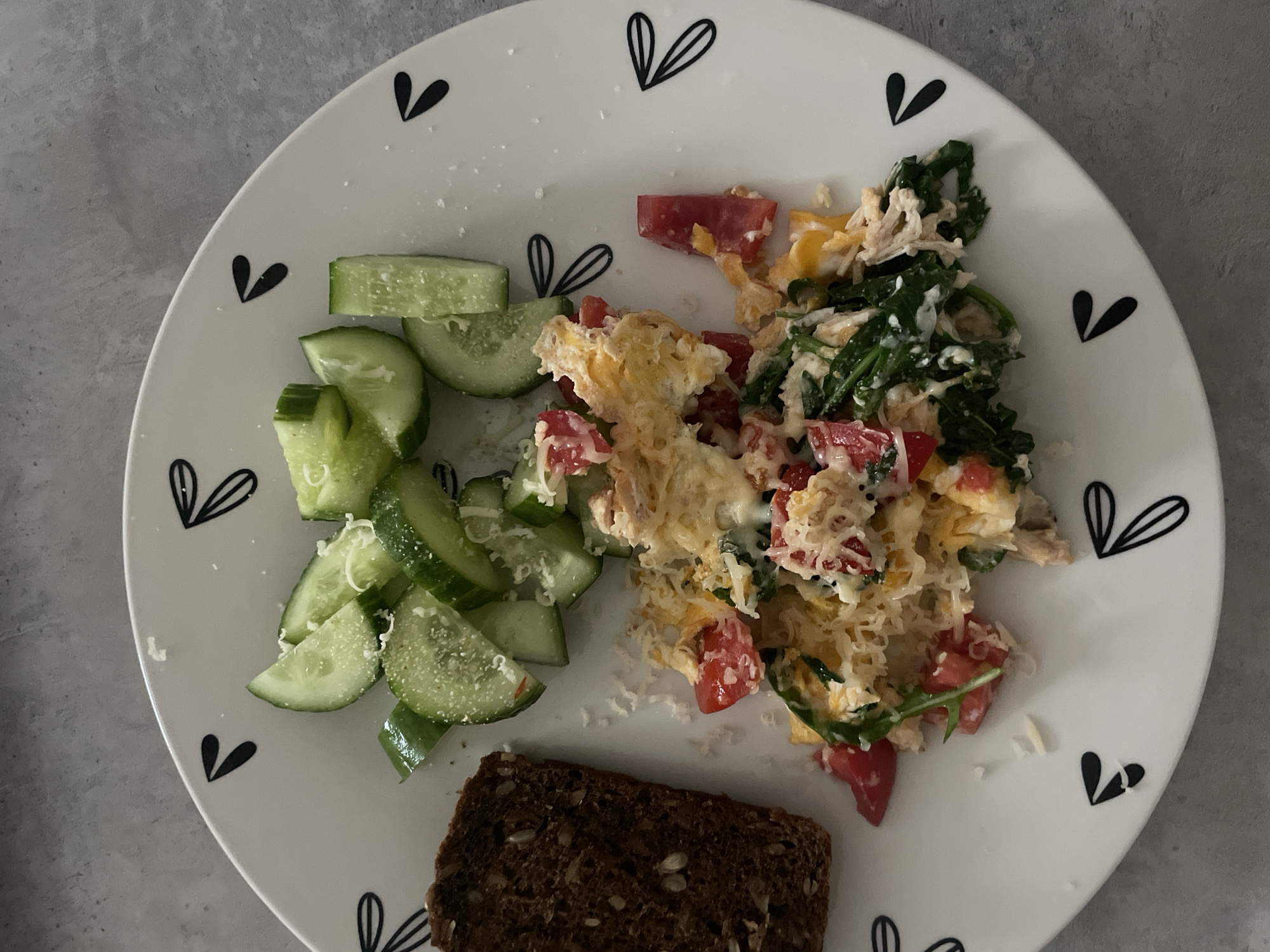 A close-up, overhead shot shows a breakfast plate on a grey, textured surface. The plate is white with a black heart-shaped leaf pattern around the rim. On the plate, there is a portion of scrambled eggs mixed with chopped tomatoes, spinach, and shredded cheese. To the left of the eggs, there are several slices of cucumber, also sprinkled with cheese. A slice of dark, seedy bread sits to the left of the cucumbers. The scene is likely a home kitchen in Poroshkino, Russia, given the context.