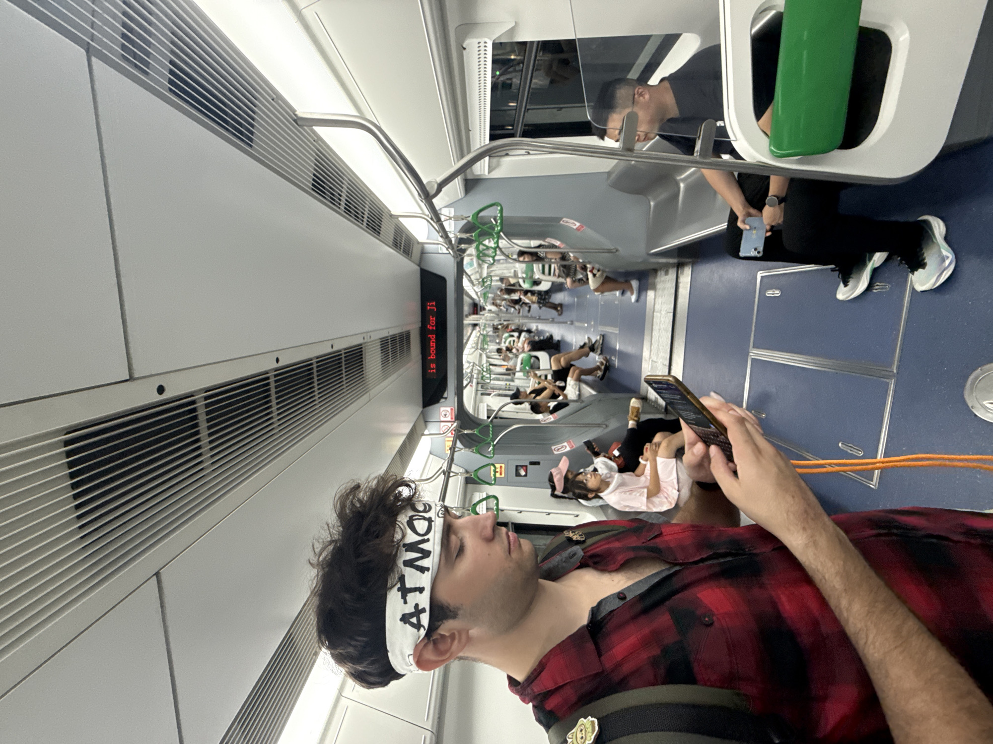 In the Yuzhong District of China, on a subway train, a man with dark, curly hair wears a white bandana with black lettering that reads "ATMOG." He is dressed in a red and black plaid shirt and holds a smartphone in his right hand. To his left, an orange lanyard hangs from his phone. In the background, other passengers are seated on the train. A young girl in a pink outfit sits across the aisle, looking down. Further down the train car, several individuals are visible, including a man with glasses in the foreground and other passengers on seats. The train's interior features grey seats, metal handrails, and overhead ventilation systems. A digital display above the seats shows red text that reads "is bound for Ji." The floor of the train car is blue.