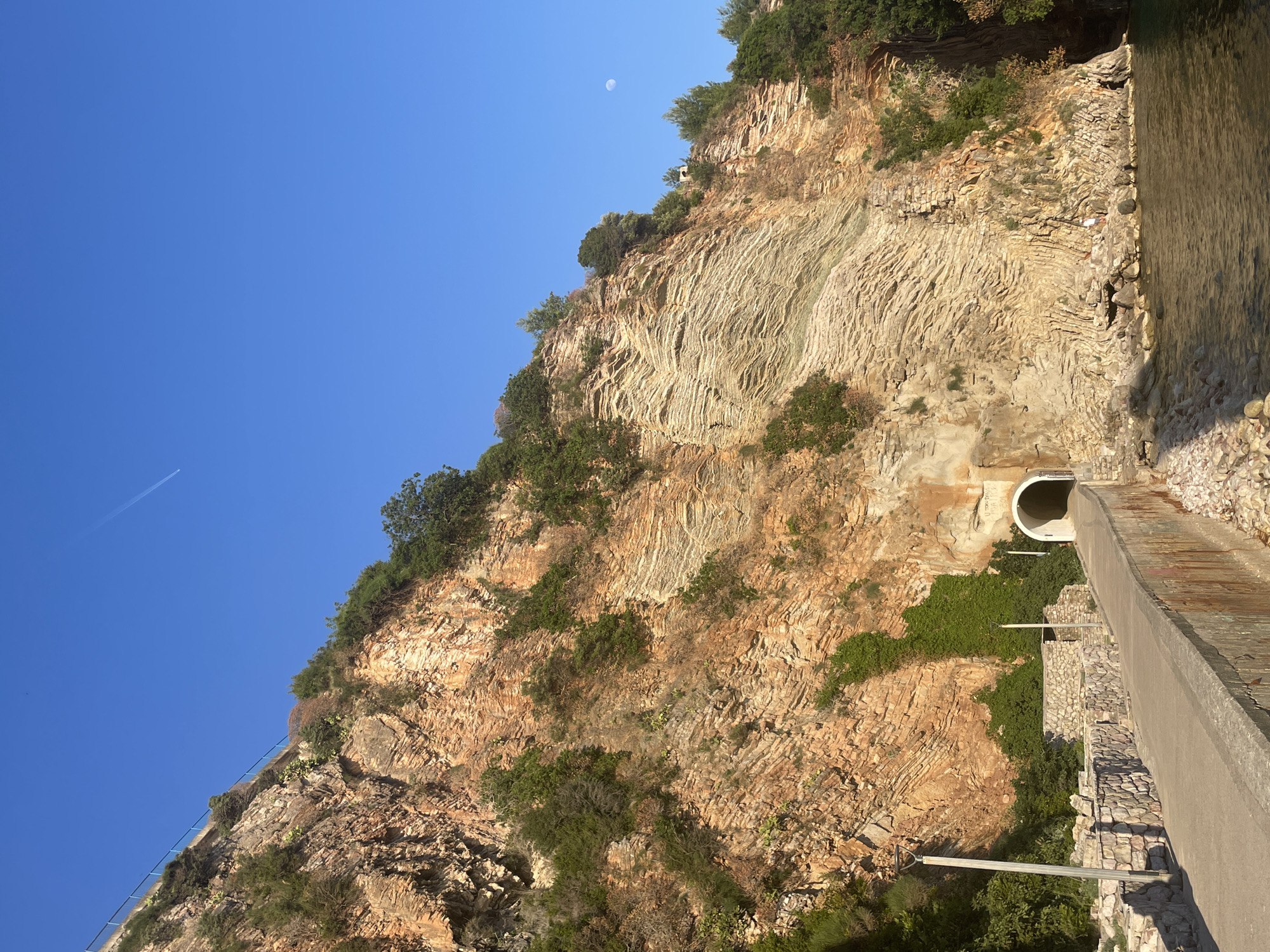 Here's a detailed description of the image:
**General Description:**
The image shows a rocky hillside in Rafailovići, Montenegro. The hillside features distinct geological strata, some vegetation, and a road with a tunnel carved into the rock.
**Specific Elements:**
* **Sky:** The sky is clear blue with a contrail from an airplane. A moon is visible.
* **Hillside:** The hillside is composed of layered rock formations. There's green vegetation growing on the hillside.
* **Road:** A gray concrete road runs along the hillside.
* **Tunnel:** A tunnel entrance is cut into the rock face and beside the road.
**Location:**
The image is taken in Rafailovići, Montenegro.
