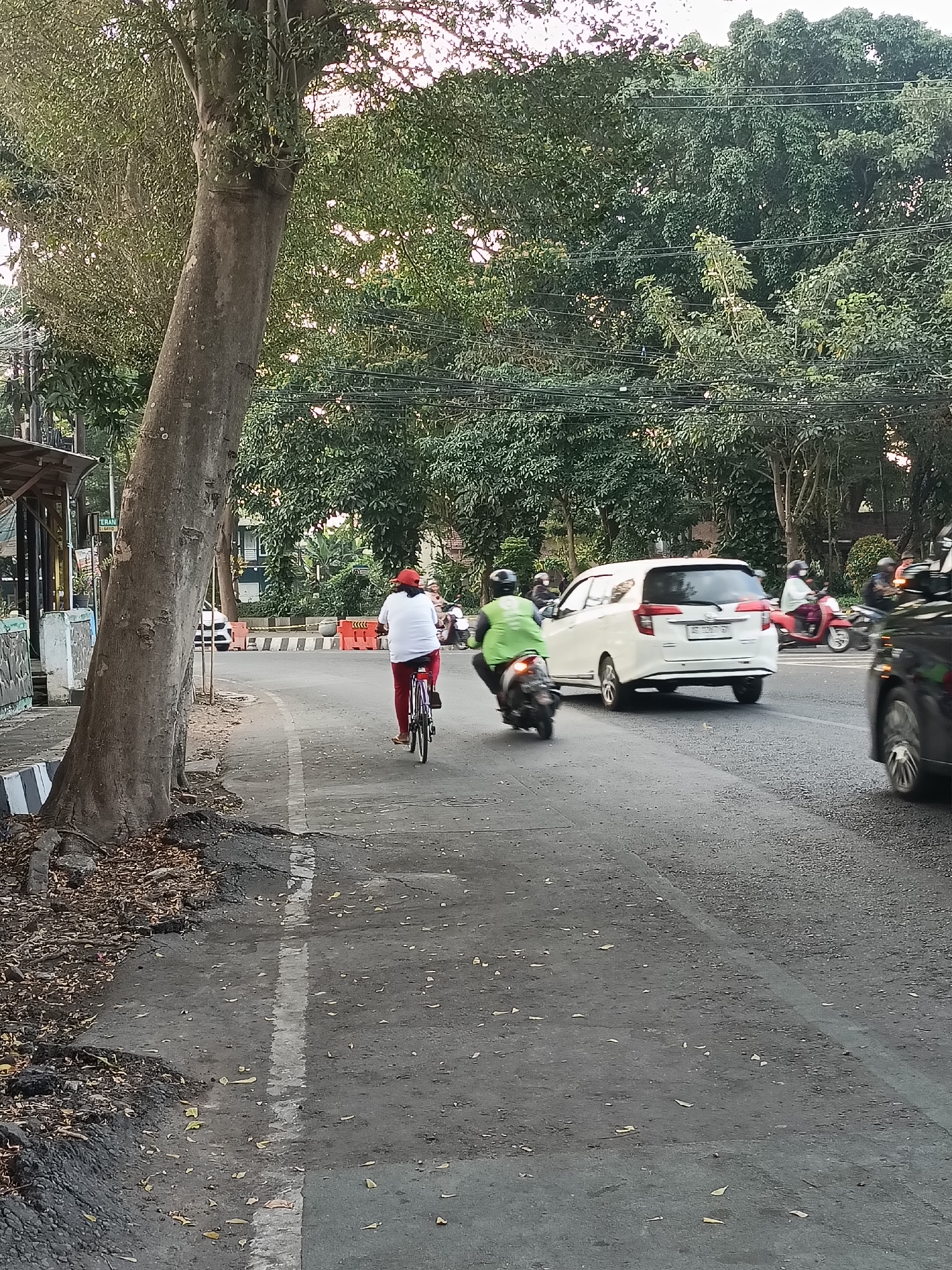 The image depicts a street scene in the city of Kediri, Indonesia, likely during daylight hours given the lighting. On the left side of the road, a person is riding a bicycle on a paved path. This individual is wearing a white shirt and red pants, and a red cap. Further down the road, a white minivan is visible, and to its right, a person on a motorcycle is riding in the same direction. In the background, dense foliage from trees lines the street, with power lines strung between them. The road surface appears to be asphalt, with some debris and fallen leaves scattered on it. The environment suggests an urban or suburban setting with significant tree cover.