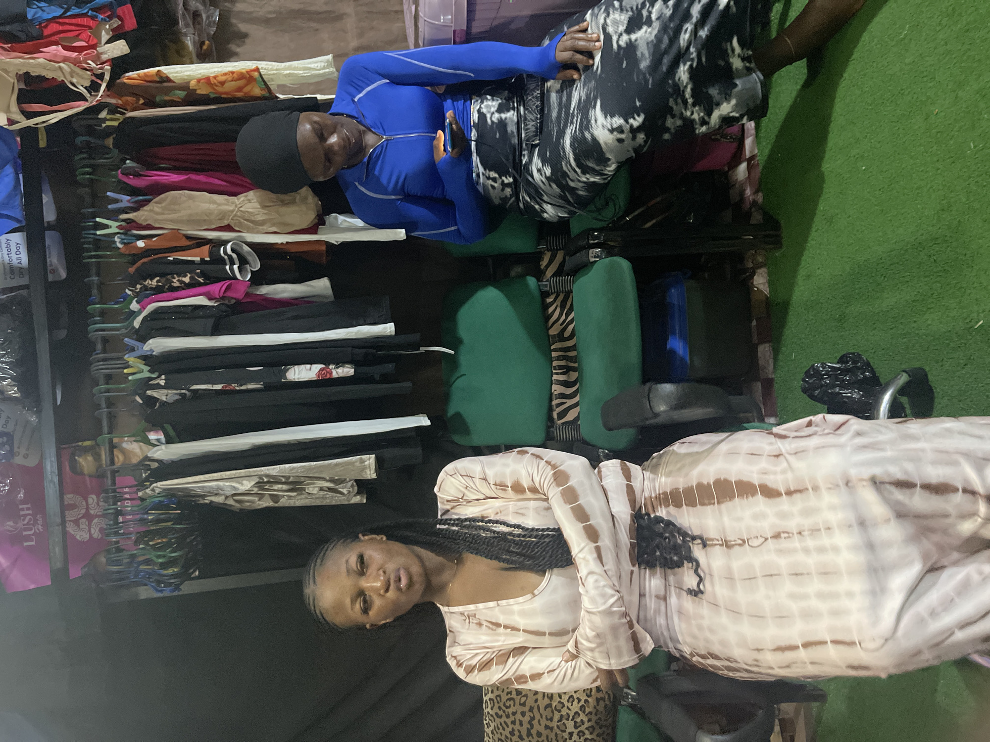 The image shows two women sitting on chairs in front of a rack of clothes. The woman on the left is wearing a light-colored, tie-dye dress and has her arms crossed. The woman on the right is wearing a blue long-sleeved top and a black and white patterned skirt. She is holding a smartphone in her hand. The environment appears to be an indoor space with green carpeting on the floor and a rack of various clothing items in the background. The scene suggests a retail or personal space where clothing is displayed or stored. There are no visible indications suggesting the location is specifically in Jos, Nigeria.