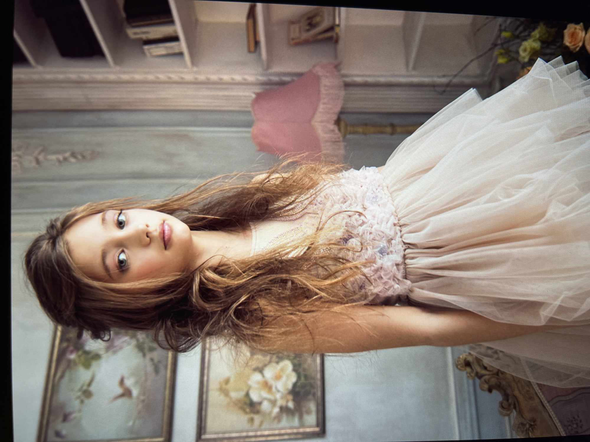 A young girl with long, wavy brown hair stands in a room with ornate furnishings and decor. She is wearing a light pink, layered dress with intricate detailing on the bodice. To her left, two framed pictures hang on the wall, one depicting a bird and the other a floral arrangement. A plush armchair with gold accents is partially visible in the foreground. To her right, a pink lampshade with fringed detailing sits on a lamp. Behind the lamp, a white bookshelf is stocked with various books. The walls have a distressed, textured finish, and decorative molding is visible. The lighting is soft and diffused, creating a warm and ethereal atmosphere. The overall impression is one of elegant and whimsical charm. The scene is set in a well-appointed interior, possibly a studio or a vintage-themed room, with no clear indication of a specific city or country, let alone Unknown, Russia.