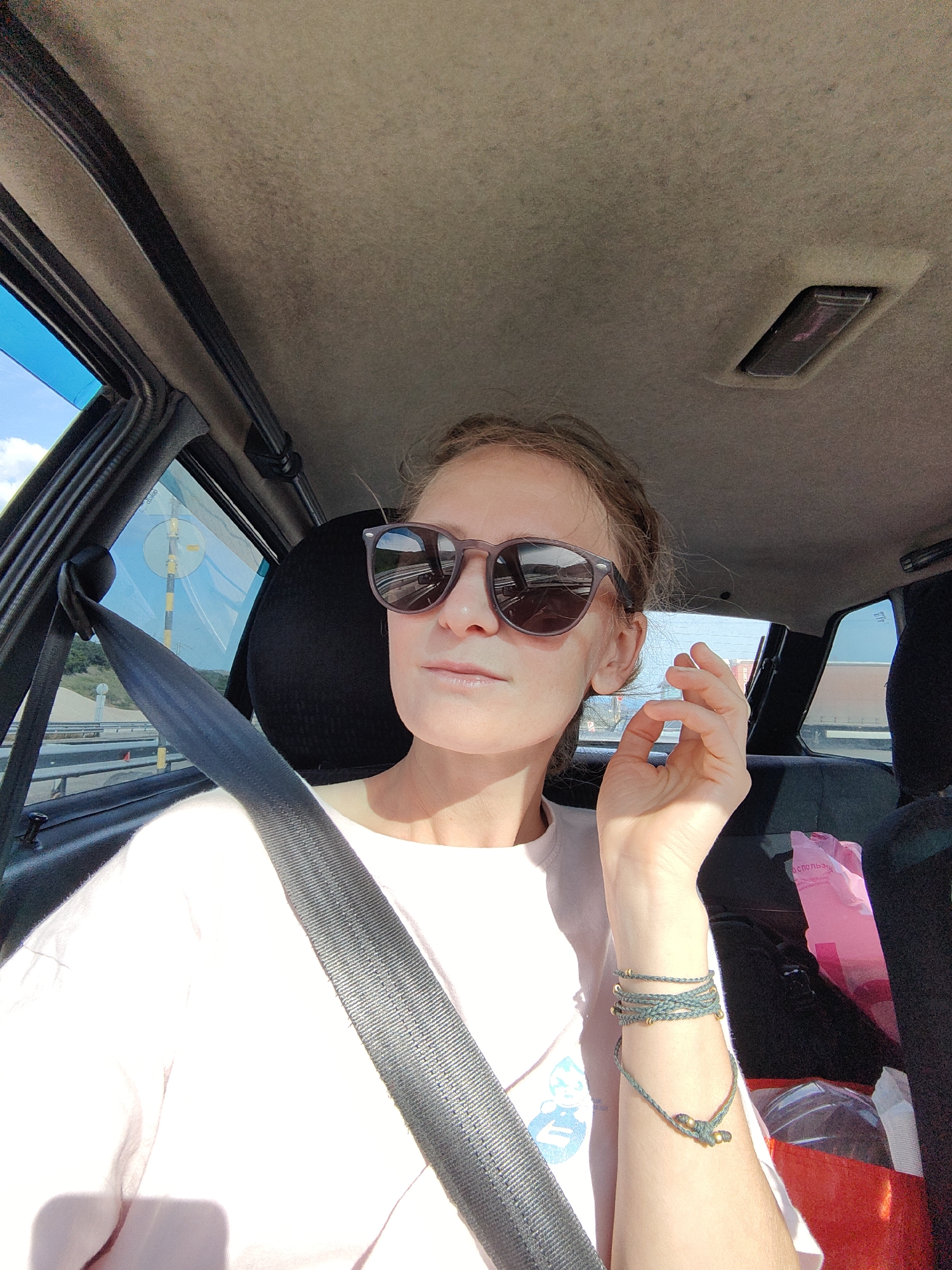 This media file shows a woman in the passenger seat of a car, taking a selfie. She is wearing sunglasses and a light-colored t-shirt. A seatbelt is fastened across her chest. Her hair is pulled back, and she has her right hand raised towards her face, with her fingers loosely arranged. She is wearing a bracelet on her left wrist.
The interior of the car is visible, including the ceiling, windows, and seats. Outside the window, a road and some greenery can be seen. The lighting suggests it is daytime, likely sunny. The overall mood appears casual and relaxed. There is no visible text.