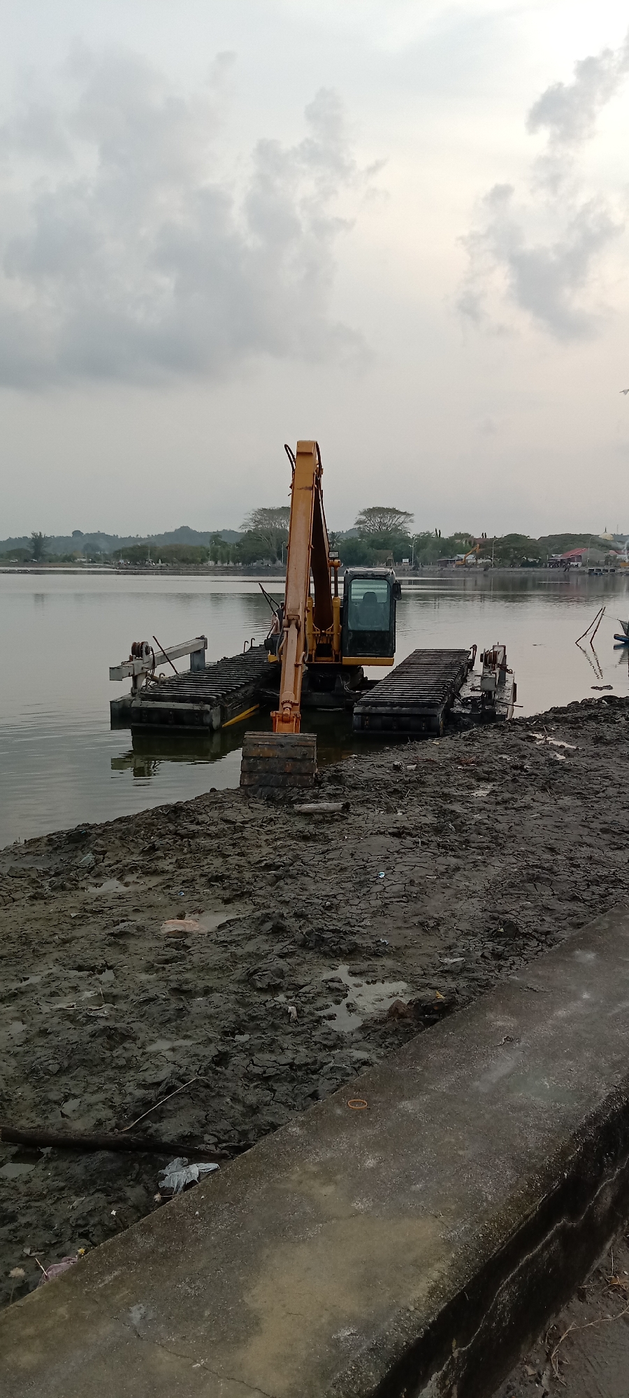 An excavator is positioned in the water near a muddy bank. The excavator sits on a platform that appears to be floating. In the background, there is a body of water with land visible in the distance. The sky is overcast. The location is likely near a waterway in Lhokseumawe, Indonesia. The muddy bank suggests recent dredging or construction activity. There are no people visible in the image.