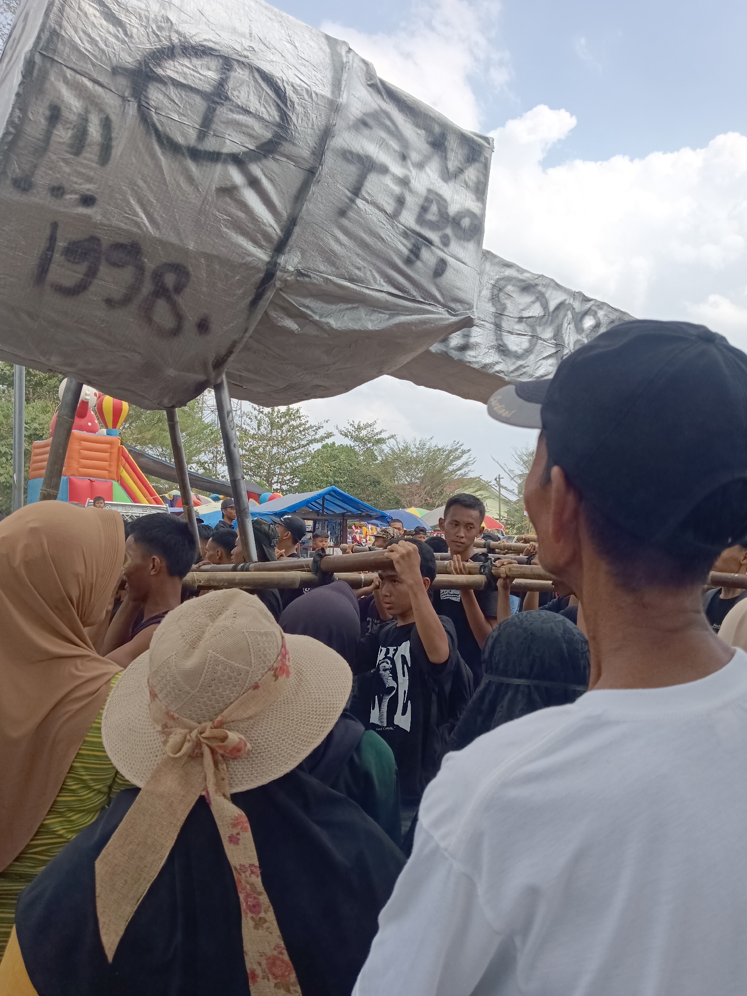 In the city of Tegal Gondo, Indonesia, a group of people are gathered outdoors under a makeshift canopy. The canopy is constructed from a large, grey tarp stretched over poles, with black spray-painted markings on it, including the year "1998." Several individuals are visible, some wearing hats, including a straw hat with a floral ribbon. Many are dressed in casual clothing, with some wearing black shirts. They appear to be participating in some sort of procession or event, as many are holding wooden poles that support the canopy. In the background, an inflatable playground structure is visible, along with other tents and trees, suggesting a public gathering or festival. The sky is partly cloudy.