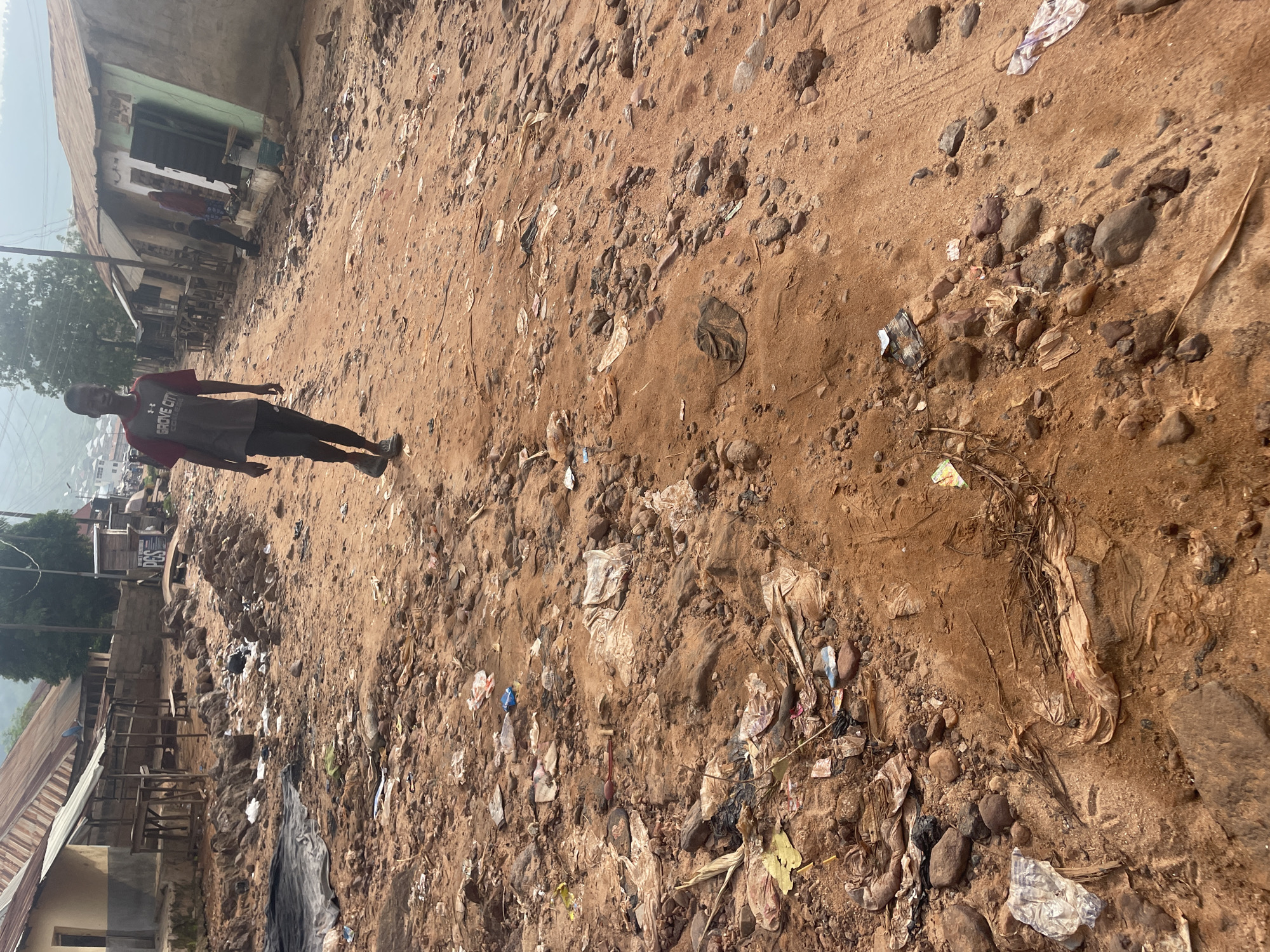 In the city of Lokoja, Nigeria, a young person walks along a dirt road littered with debris and trash. The road is unpaved and appears to be in a state of disrepair. Buildings line the sides of the road, some with corrugated metal roofs. The overall environment suggests a developing urban area with visible signs of waste management challenges. The young person is wearing a t-shirt and pants, walking away from the camera and towards the background. The ground is a mix of reddish-brown dirt, rocks, and various forms of litter, including plastic bags and wrappers. The scene is set during daytime, with natural light illuminating the area.