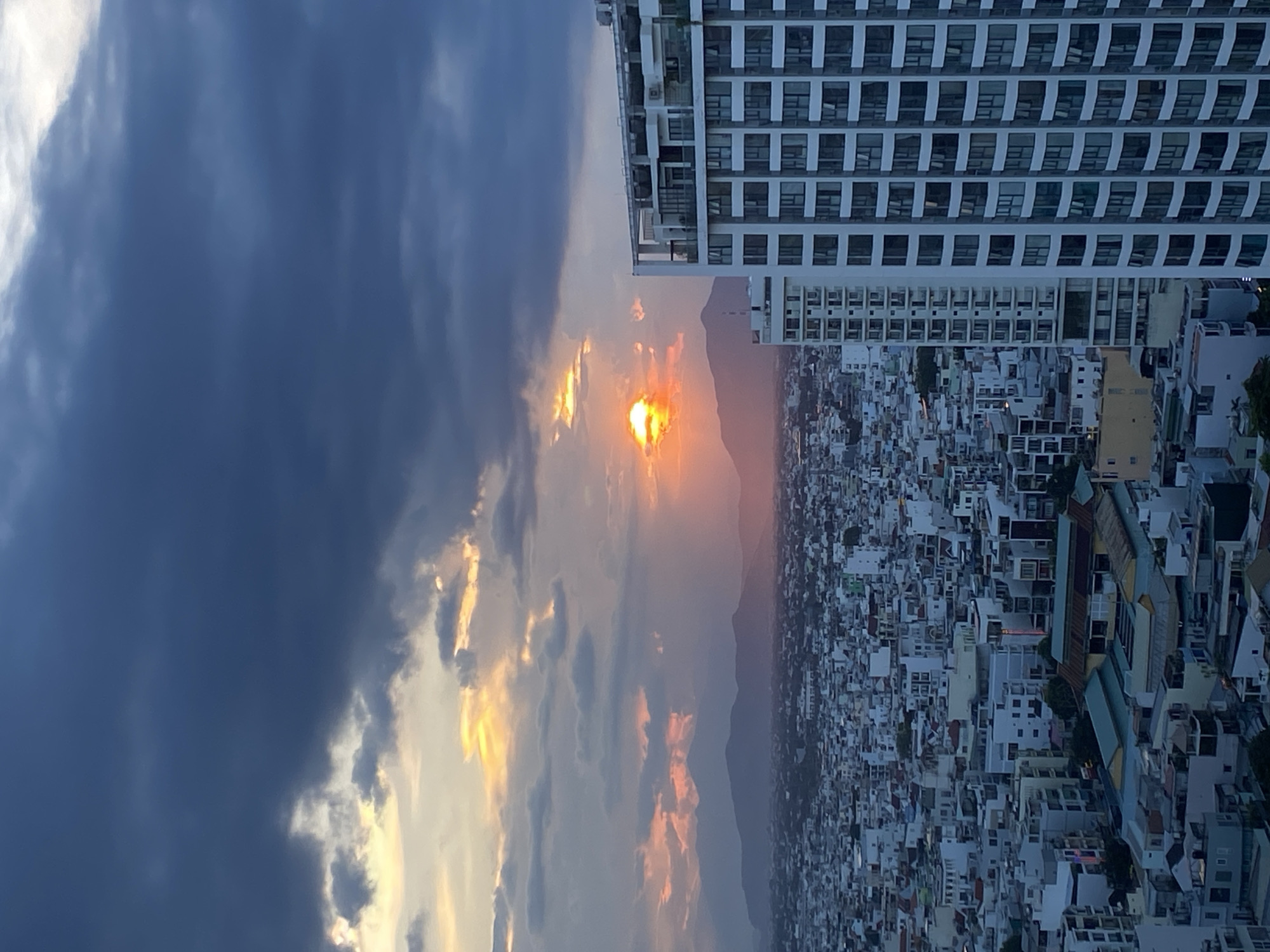 This is an aerial view captured during sunset in Nha Trang Ward, Vietnam. The sky is filled with dramatic, dark clouds that are partially illuminated by the setting sun, which appears as a bright orange orb with yellow and pinkish hues filtering through the clouds and reflecting on the mountains in the distance. A vast cityscape stretches out below, densely packed with buildings of varying sizes and architectural styles, predominantly white, grey, and some with colored rooftops. In the foreground, a tall, modern white building with numerous windows dominates the right side of the frame. To the left of this building, another, shorter white building is partially visible, and beyond that, the dense urban sprawl continues towards a range of dark, silhouetted mountains that form the horizon. The overall impression is of an urban landscape at dusk, with the natural beauty of the sunset contrasting with the man-made environment. No people are visible in the image.
