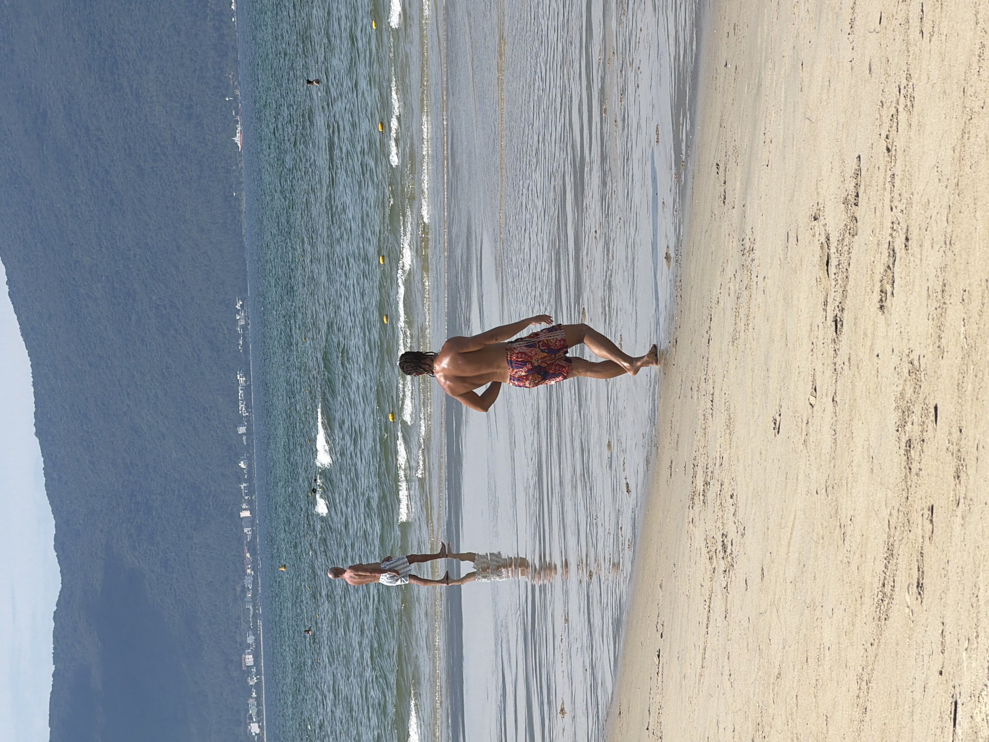 In Đà Nẵng, Vietnam, on a sandy beach, two men are walking near the ocean. The man in the foreground, with dark, curly hair, is walking away from the camera, wearing patterned swim trunks. The man in the background, bald, is walking towards the right, also in swim trunks. The ocean is calm with small waves and yellow buoys marking a designated swimming area. In the distance, a lush green mountain range rises behind a line of buildings, suggesting a coastal city setting. The sky is partly cloudy, and the sun casts reflections on the wet sand.