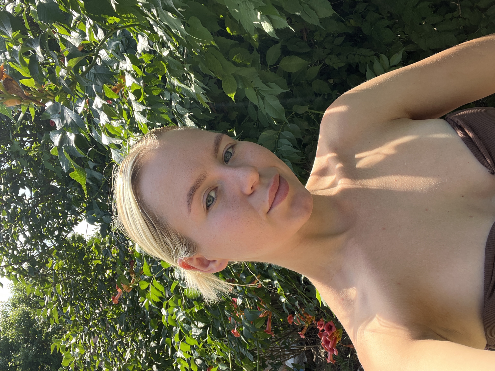 An outdoor, eye-level, medium shot captures a young Caucasian woman with blonde hair pulled back, facing the camera with her head tilted slightly. She wears a brown strapless top and is positioned in front of a dense green foliage background with some red flowers visible on the left. The lighting suggests it is daytime, with shadows cast across the woman's shoulder and arm, indicating the sun is to her right. Her skin appears slightly flushed and dewy, possibly from sun exposure. Her facial expression is neutral, with a subtle smile. The background is out of focus, emphasizing the woman as the central subject.