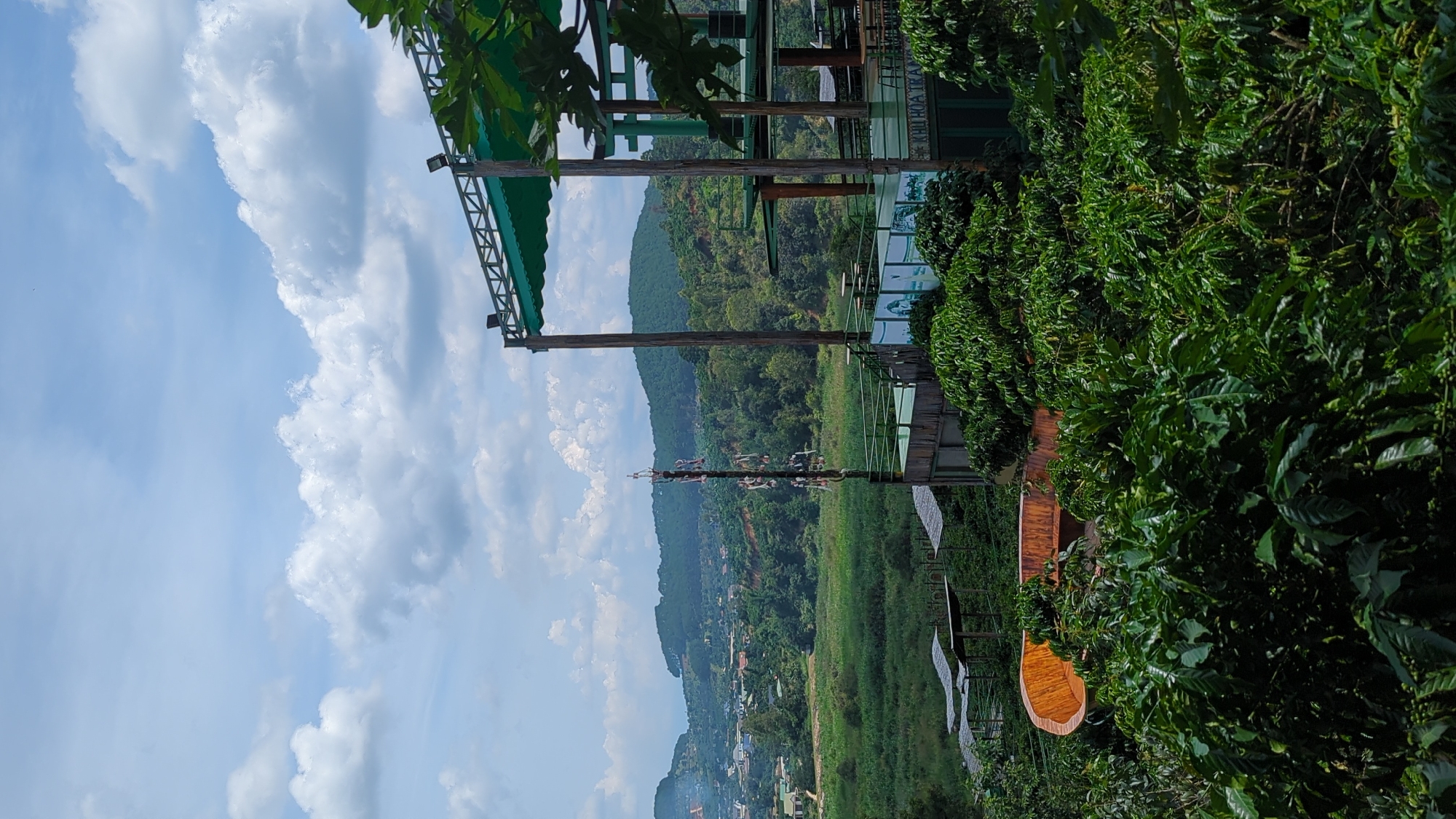The image depicts an outdoor scene with a layered landscape and a multi-level structure in the foreground. A lush green forest dominates the midground, with rolling hills and mountains in the background. The sky is partly cloudy with a mix of white clouds and blue sky.
In the foreground, a large, wooden, boat-shaped seating area is partially visible amidst dense green foliage. To the right, a multi-story building with a green awning and wooden pillars is situated on a raised platform, with tables and chairs visible. A cell tower stands tall to the left of the building. The terrain appears to be hilly and overgrown with vegetation.
The scene is situated in Thôn 5, Vietnam, a region known for its natural beauty and agricultural landscapes. The environment suggests a rural or semi-rural setting, possibly a tourist attraction or a viewpoint overlooking the countryside. The lighting indicates it is daytime, with the sun illuminating the scene, creating some shadows. No people are visible in the image.