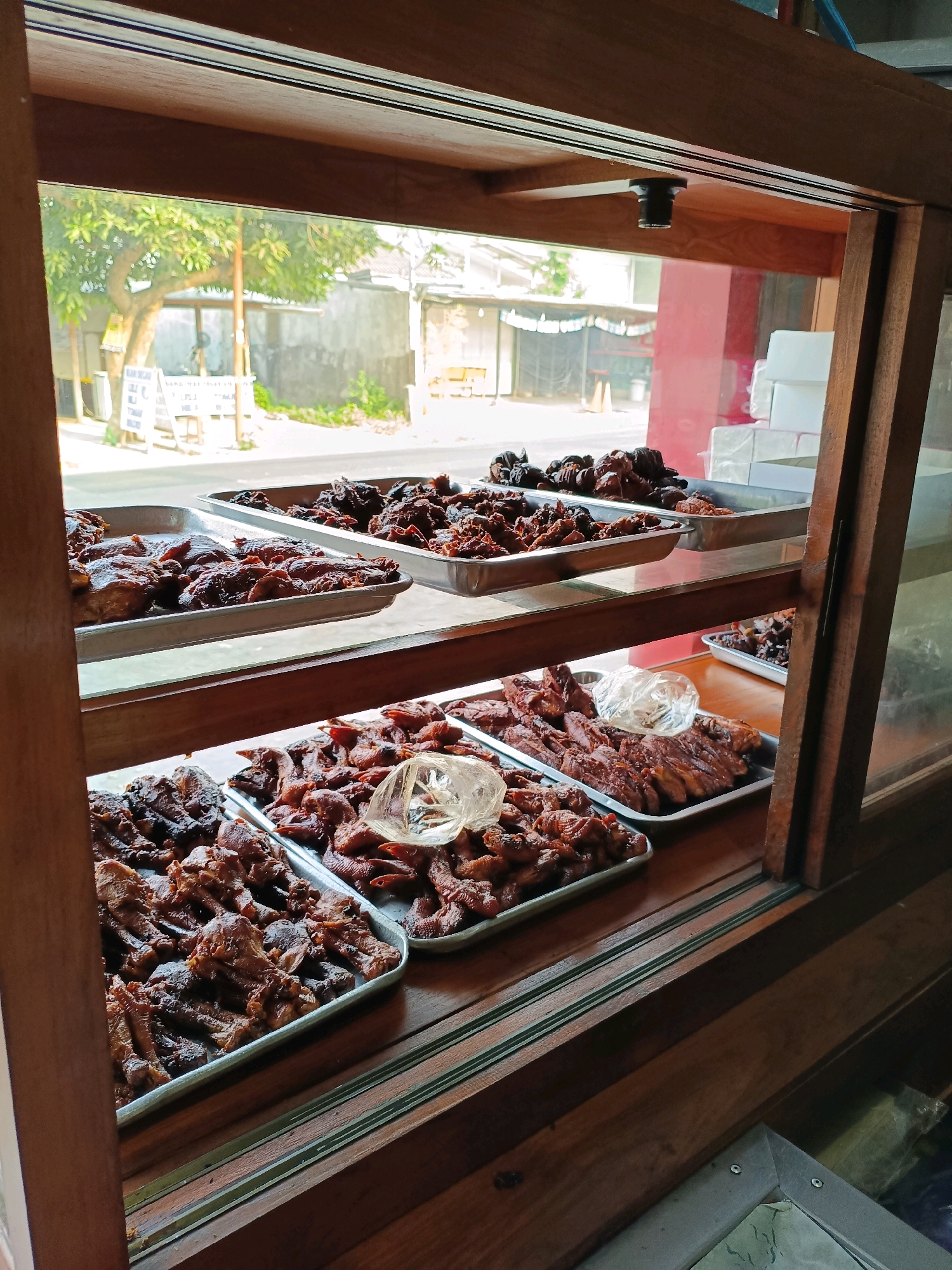 This is a photograph taken at a food stall in Maguwoharjo, Indonesia. The image is taken from outside looking in through a glass display case, which is framed by dark wood. Inside the case, there are multiple trays of cooked food, primarily consisting of various cuts of meat, appearing to be chicken, that are dark brown and glazed, suggesting they have been fried or roasted. The food is arranged in shallow metal trays, stacked on wooden shelves.
In the background, visible through the glass, is an outdoor scene. There are trees with green foliage, a street, buildings with neutral-colored walls, and a red wall on the right side of the frame. There is also a white sign with text on it. The lighting suggests it is daytime, with natural light illuminating the scene. No people are directly visible interacting with the food. The overall impression is of a street-side food vendor displaying its offerings.