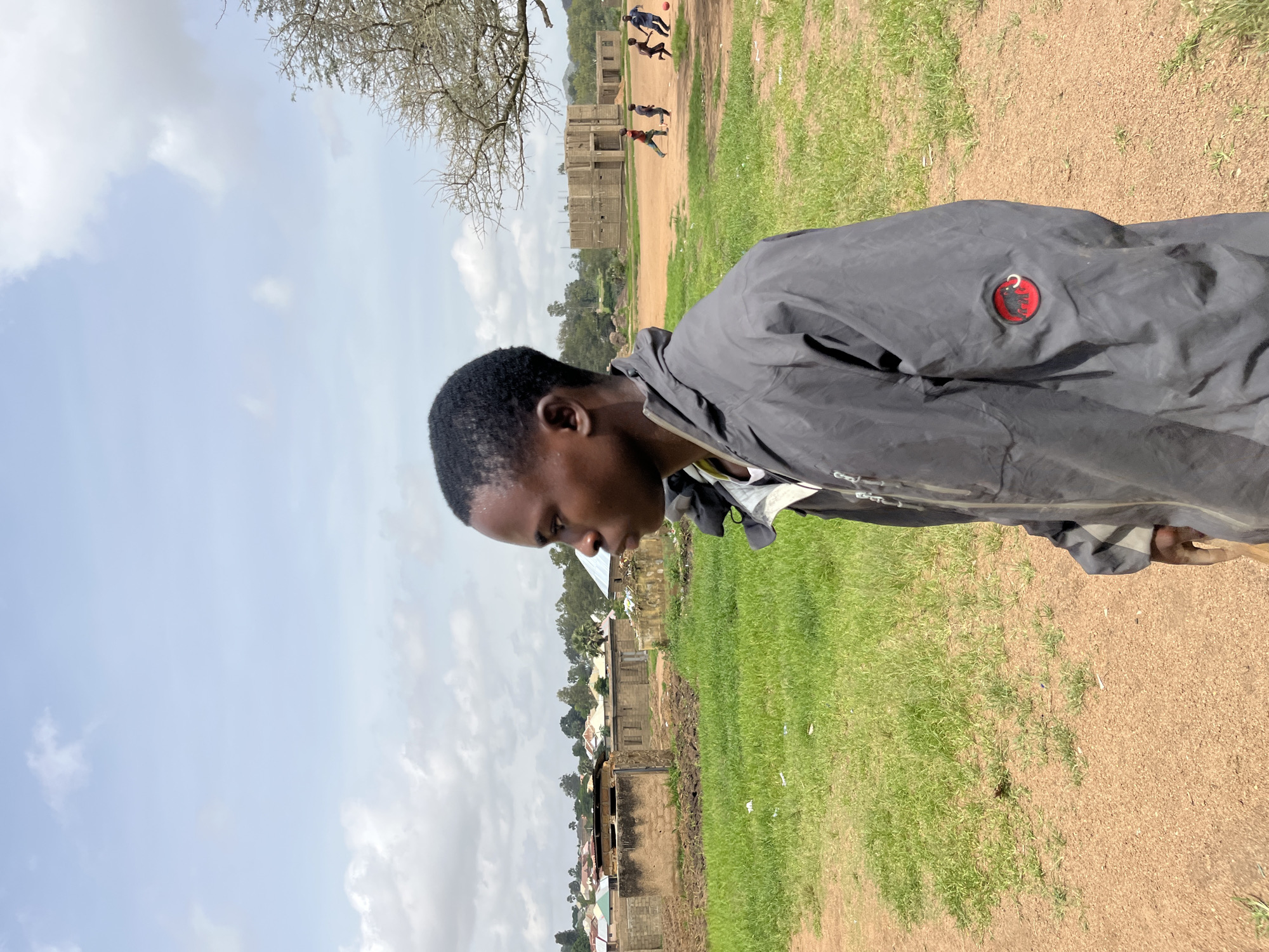 The image depicts a young Black male in the foreground, facing right in profile, wearing a dark grey hooded jacket with a red circular emblem on the left sleeve. He has short, dark hair and is looking off into the distance. The background reveals an outdoor scene that appears to be within the city of Bukuru, Nigeria. In the middle ground, there are several structures, some appearing to be made of mud brick, with corrugated metal roofs. Some of these buildings seem incomplete or in a state of disrepair. Further in the background, more houses with colorful roofs are visible, nestled amongst trees. To the right of the image, a group of children are playing soccer in a dusty, open field. A bare-branched tree stands prominently on the right side of the frame. The ground in the immediate foreground is a reddish-brown dirt, transitioning to green grass in the middle ground. The sky is partly cloudy, with bright white clouds against a pale blue backdrop. The overall impression is a snapshot of everyday life in a Nigerian community.